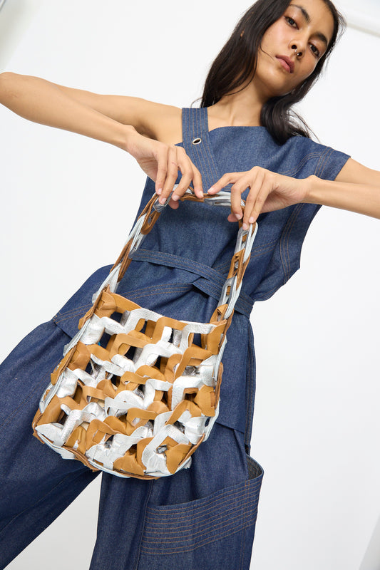 A woman in a blue denim outfit lifts her arms to display the SC103 Leather Links Tote in Parcel, a unique handmade bag crafted from tan and silver materials.