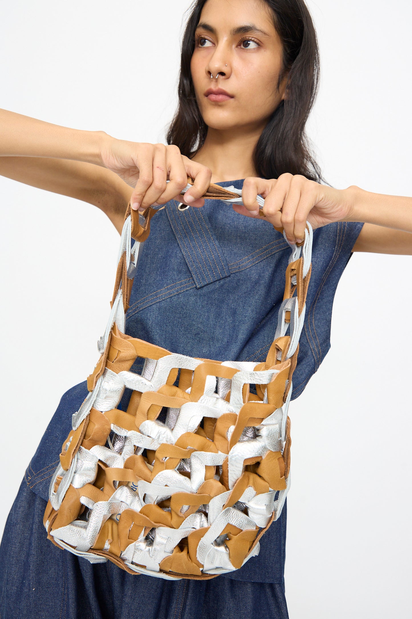 A person in a blue denim dress holds the SC103 Leather Links Tote in Parcel—handmade with interlocking metallic silver and tan leather strips—against a plain white background.