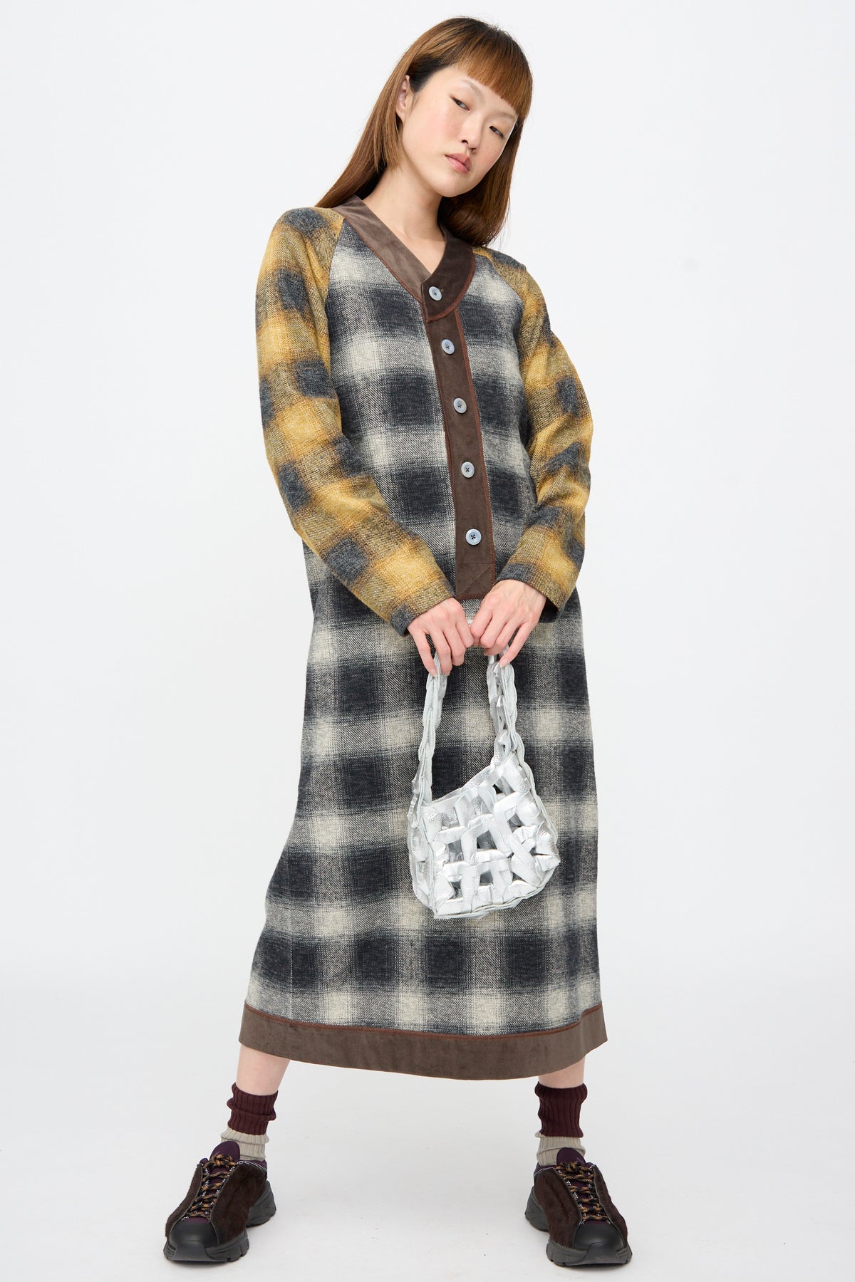 A person wearing a long, plaid, button-front dress and dark sneakers, holding a silver, woven Leather Links Tote Mini in Glow.