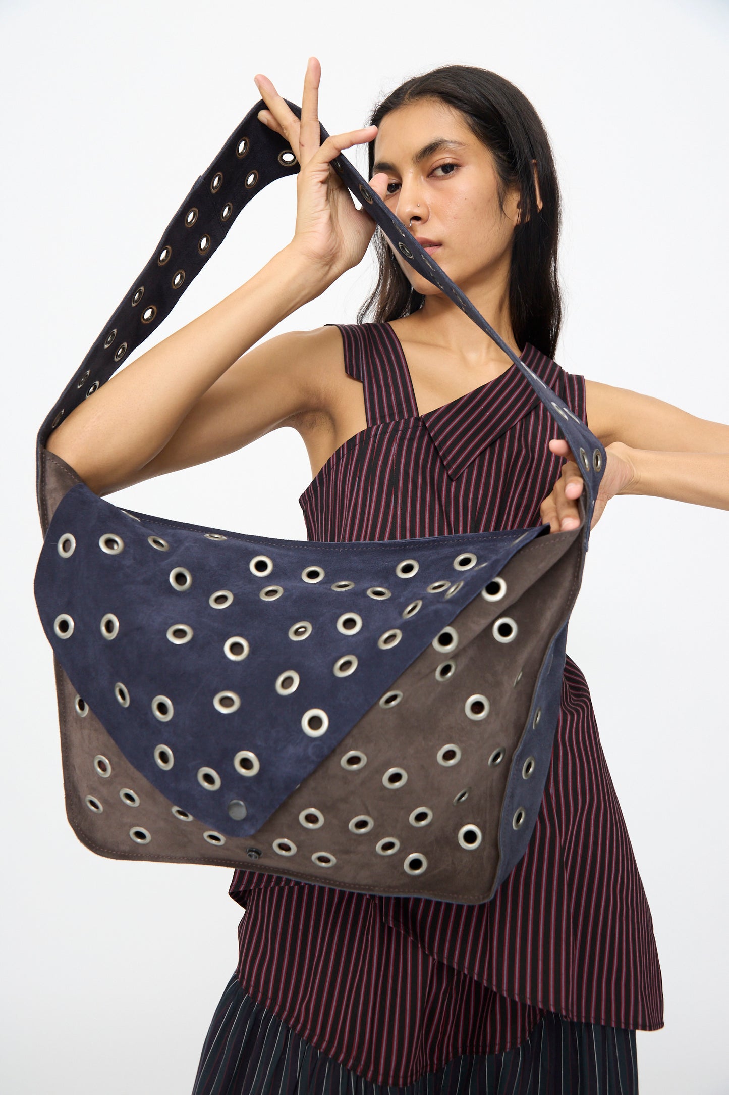 A woman in a striped sleeveless top holds the SC103 Leather Suede Record Messenger Bag in Moonspell, featuring a blue and brown geometric pattern with grommets and suede patchwork accents.