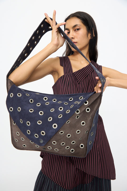 A woman in a striped sleeveless top holds the SC103 Leather Suede Record Messenger Bag in Moonspell, featuring a blue and brown geometric pattern with grommets and suede patchwork accents.