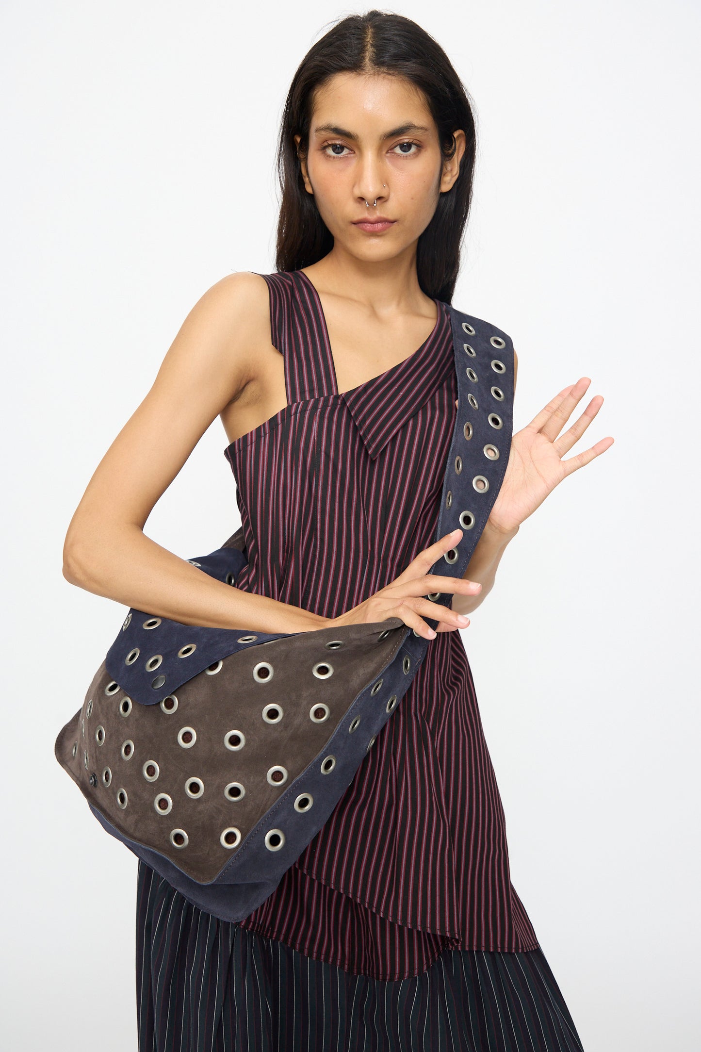 A woman with long dark hair wears a sleeveless, asymmetrical striped top and carries the SC103 Leather Suede Record Messenger Bag in Moonspell across her shoulder.