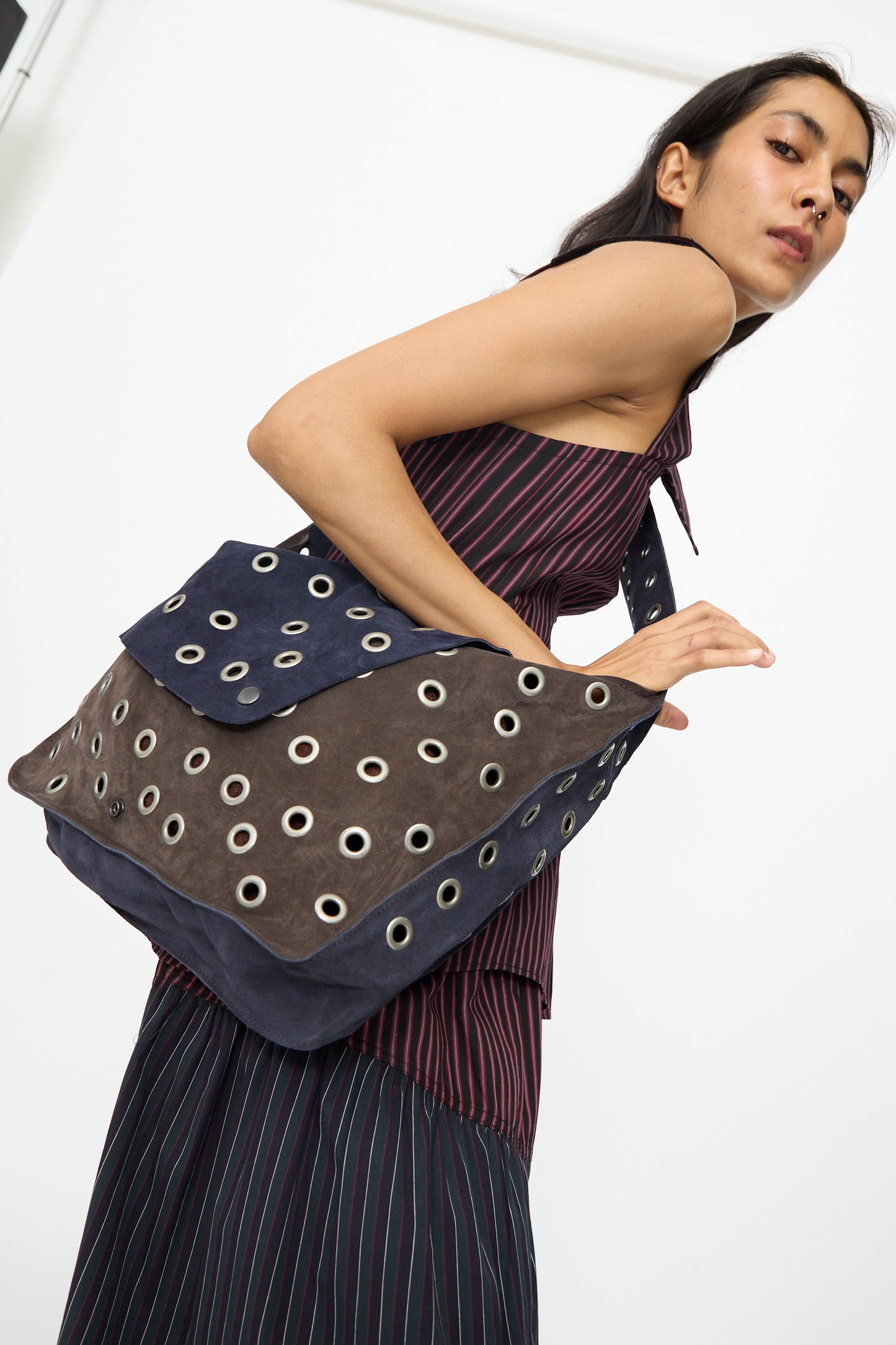 A woman in a striped dress carries the SC103 Leather Suede Record Messenger Bag in Moonspell, featuring metallic grommet details and navy and brown suede patchwork.