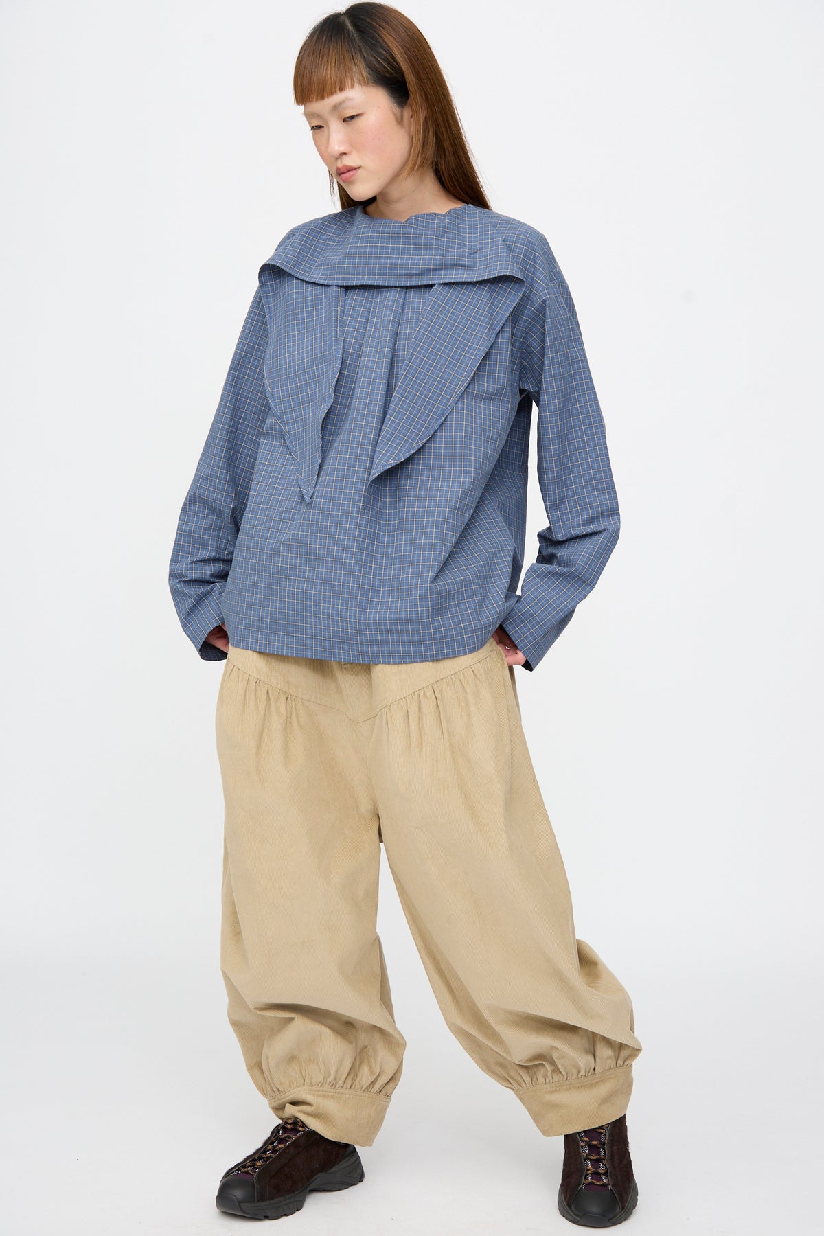 A person wears an SC103 Plaid Poplin Compass Blouse in Aerial, a blue plaid cotton long-sleeve top with the neck tie undone, paired with loose-fitting tan trousers with gathered cuffs.