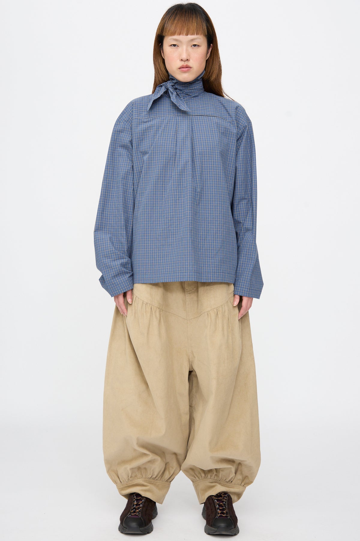 Model wearing a SC103 Plaid Poplin Compass Blouse in Aerial with a high neck tie and loose-fitting, beige balloon-style trousers.
