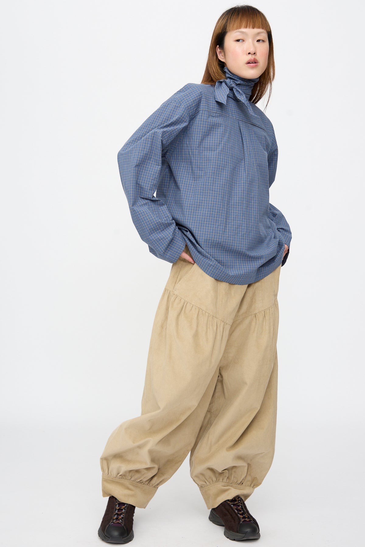 A person wearing a blue Plaid Poplin Compass Blouse by SC103 with loose-fitting tan trousers with gathered cuffs and dark sneakers.