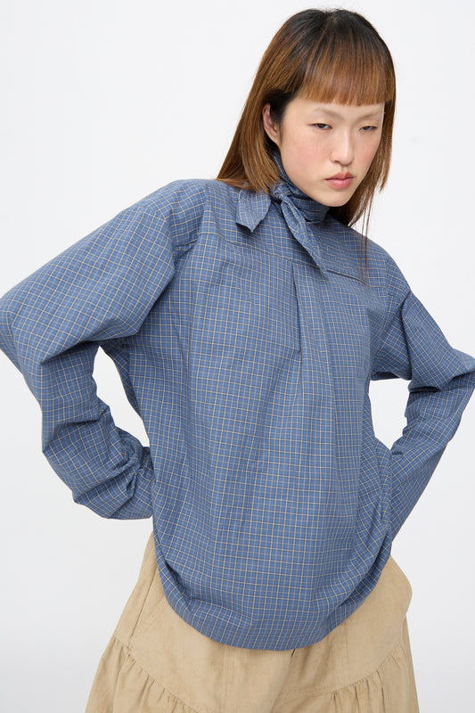 A person wearing a Plaid Poplin Compass Blouse in Aerial, a long sleeve blue plaid cotton blouse with a high neck tie, worn with tan trousers.