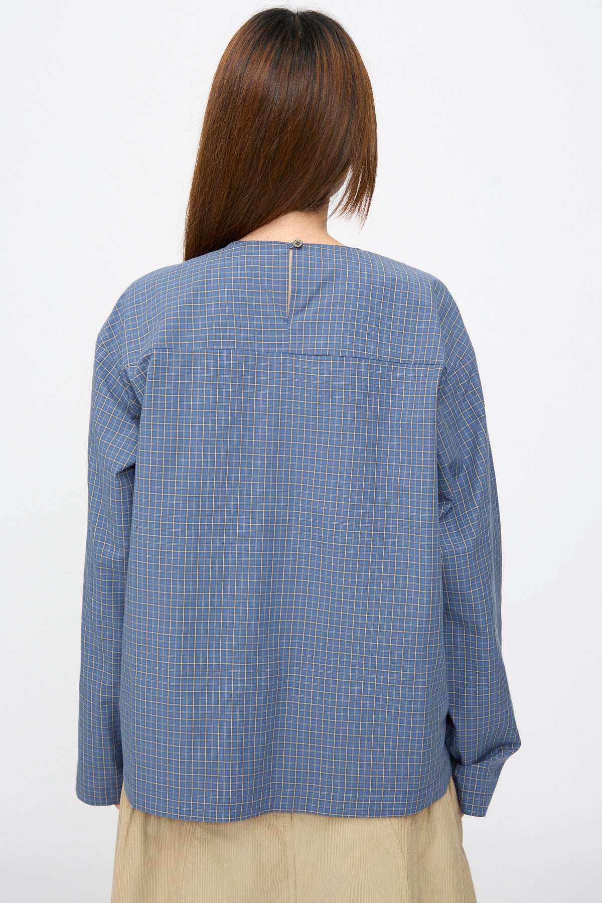 Back view of a person wearing an SC103 Plaid Poplin Compass Blouse in Aerial, featuring a blue plaid cotton long-sleeved design with a single button closure at the nape of the neck.