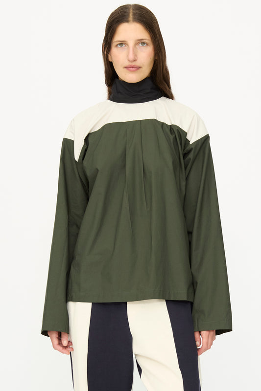 A woman with long brown hair wears the SC103 Poplin Compass Blouse in Relay, a loose-fitting, long sleeve green and cream top, paired with black and white pants against a plain white background.