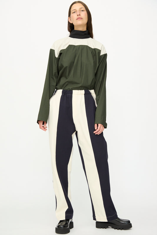 A person stands against a white background wearing SC103 Rib Knit Leap Sweats in Index—relaxed fit cotton knit pants with bold vertical navy and white stripes—paired with a color-blocked long-sleeve top and black platform shoes. Made in NYC.