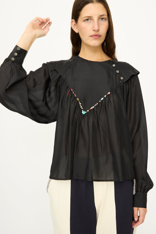 A woman wears the SC103 Silk Prince Blouse in Thunder— a loose, black, long-sleeve silk blouse with buttons and beaded details—styled with black and white color-blocked pants, standing against a plain background.
