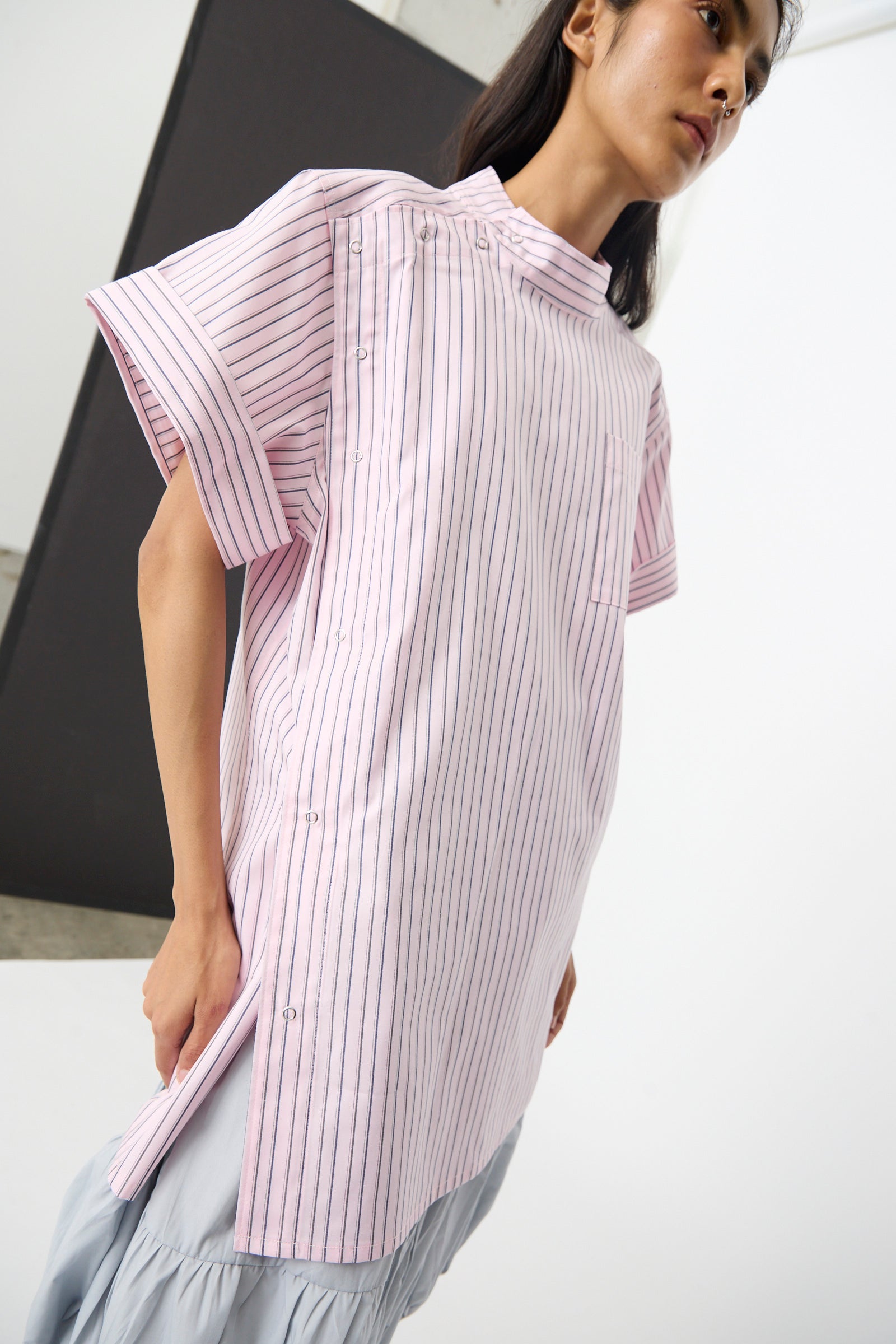 A person wearing the SC103 Stripe Cotton Wish Top in Blushline, featuring pink and white stripes, a band collar, short sleeves, and side slits, stands against a minimal background.
