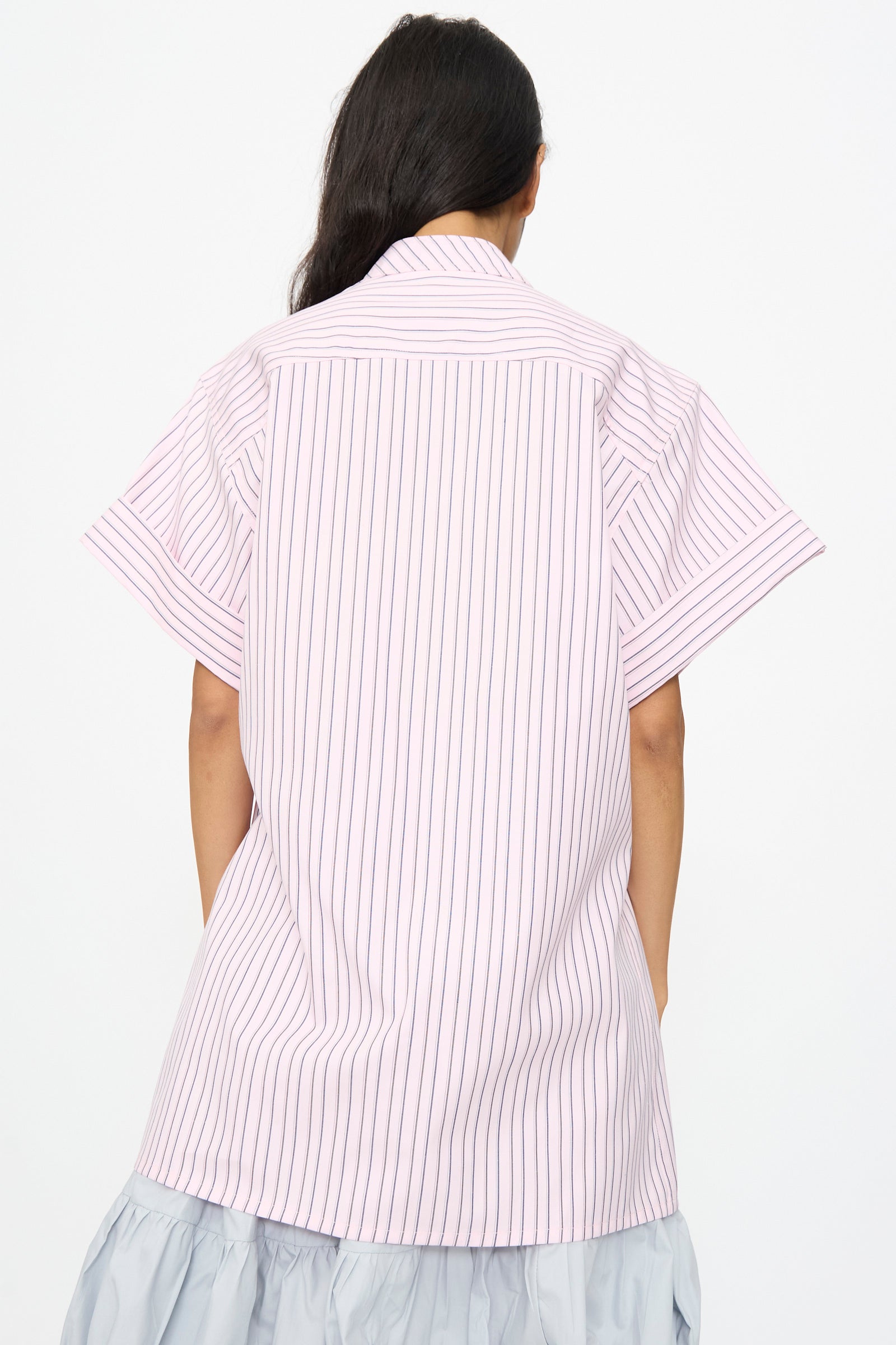 A person faces away, wearing the SC103 Stripe Cotton Wish Top in Blushline, a light pink short-sleeve shirt with vertical black stripes and a band collar, paired with a light gray skirt.