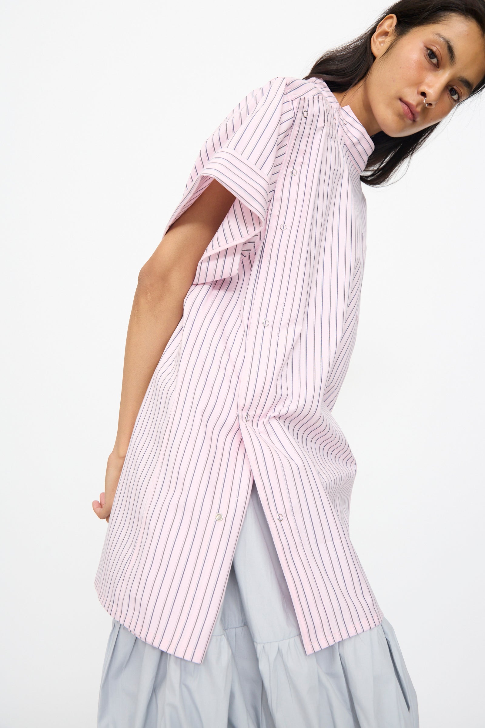 A woman models the SC103 Stripe Cotton Wish Top in Blushline—featuring a band collar, back buttons, and high slit—over a light gray skirt, standing against a plain background.