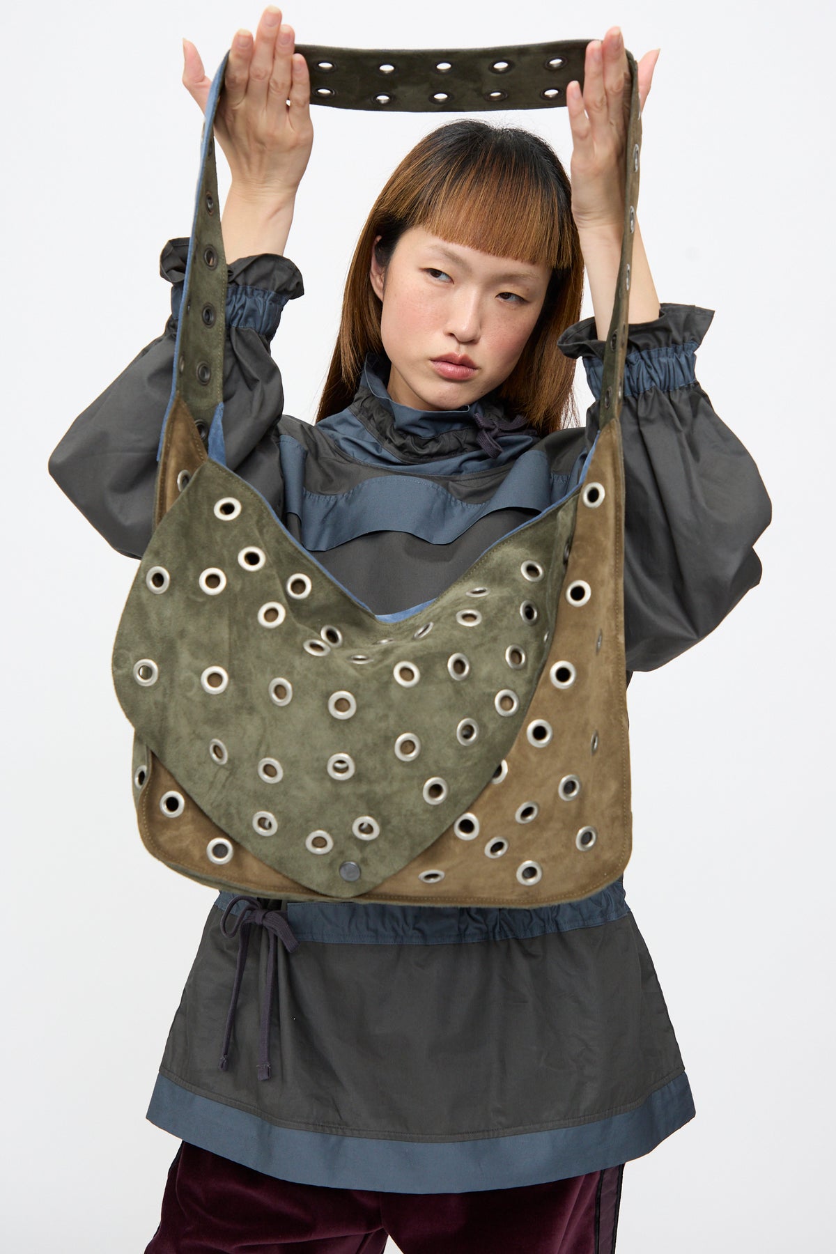 A person holds up the SC103 Suede Leather Record Messenger bag, featuring a patchwork design decorated with numerous metal grommets.