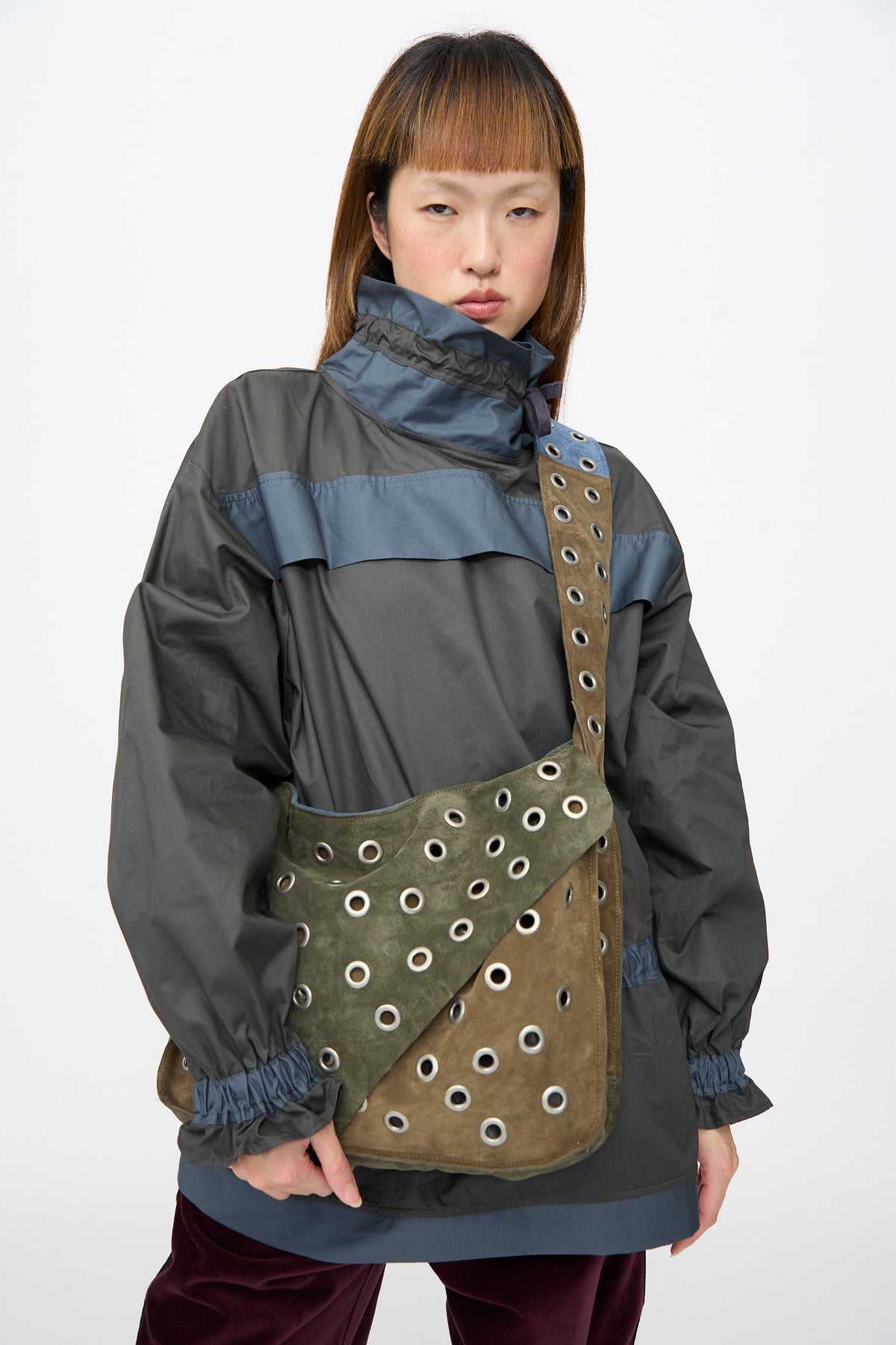 A person wearing a dark grey top and the SC103 Suede Leather Record Messenger in Abyss, a patchwork bag with a wide strap featuring multiple metal grommets.