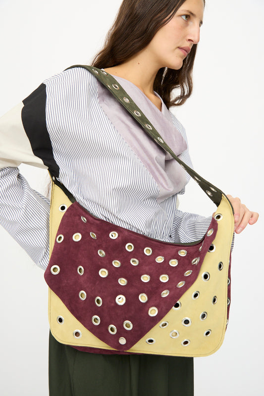A woman wearing a striped blouse carries the SC103 Suede Record Messenger in Campus, a large maroon and beige patchwork shoulder bag with metal grommets.