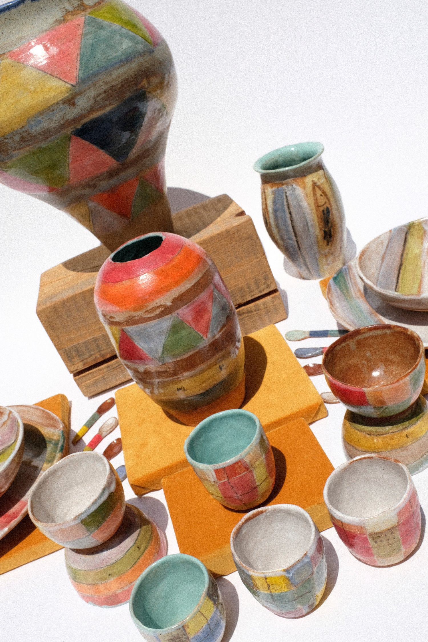 A collection of handmade ceramic tableware, including a large vase, a small jar, two tall narrow vases, bowls, and cups, all decorated with colorful geometric patchwork patterns in orange, red, green, yellow, blue, and brown, displayed on wooden blocks against a white background.