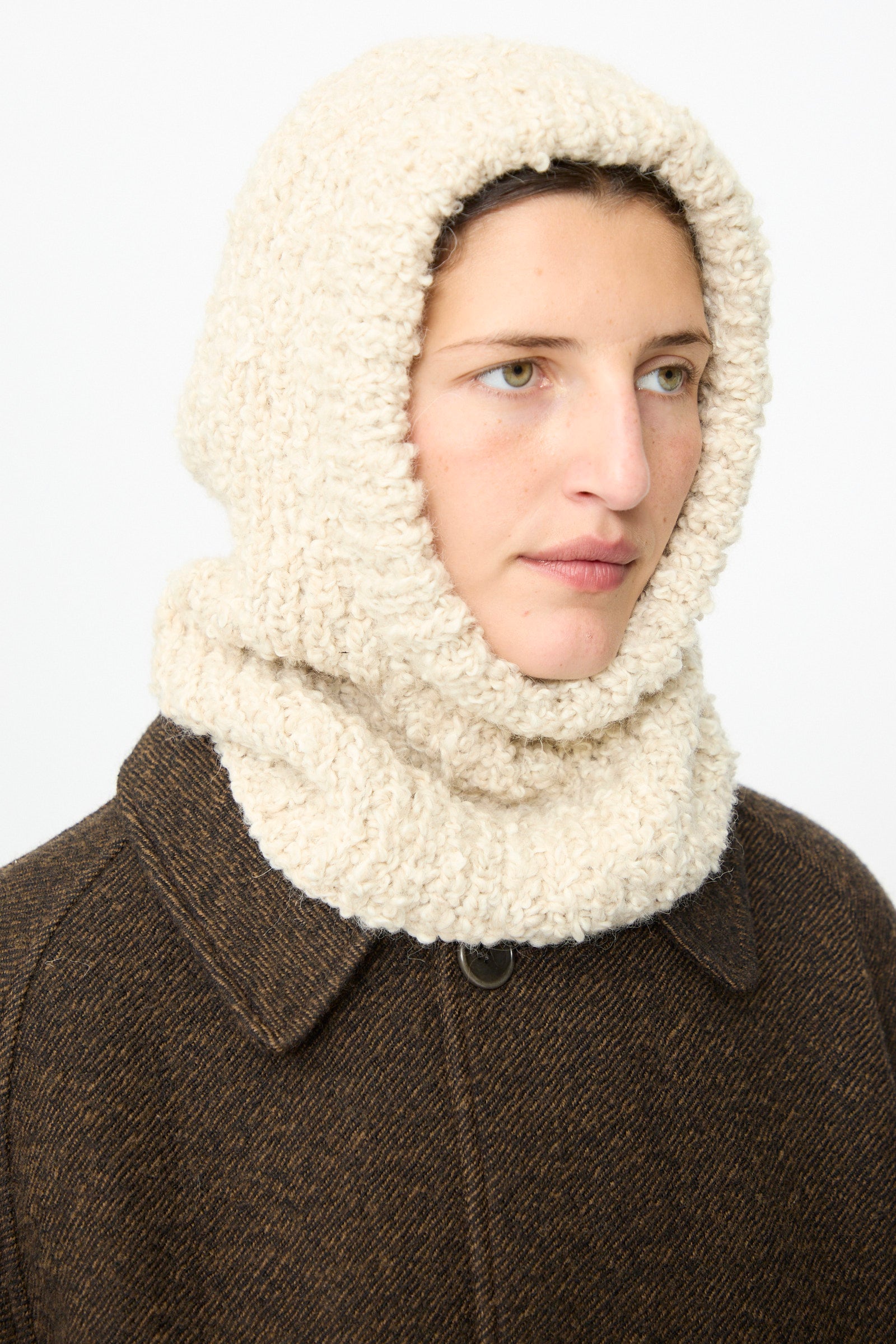 A person wearing a brown coat and the SONO Bruna Balaclava in Natural—a cream-colored, ribbed wool-alpaca blend hooded scarf—looks slightly to the right against a plain background.