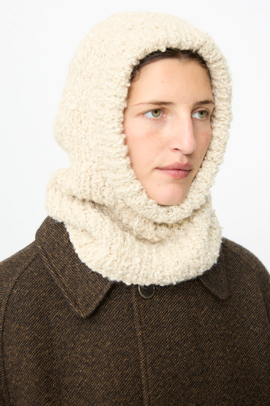 A person wearing a brown coat and the SONO Bruna Balaclava in Natural—a cream-colored, ribbed wool-alpaca blend hooded scarf—looks slightly to the right against a plain background.