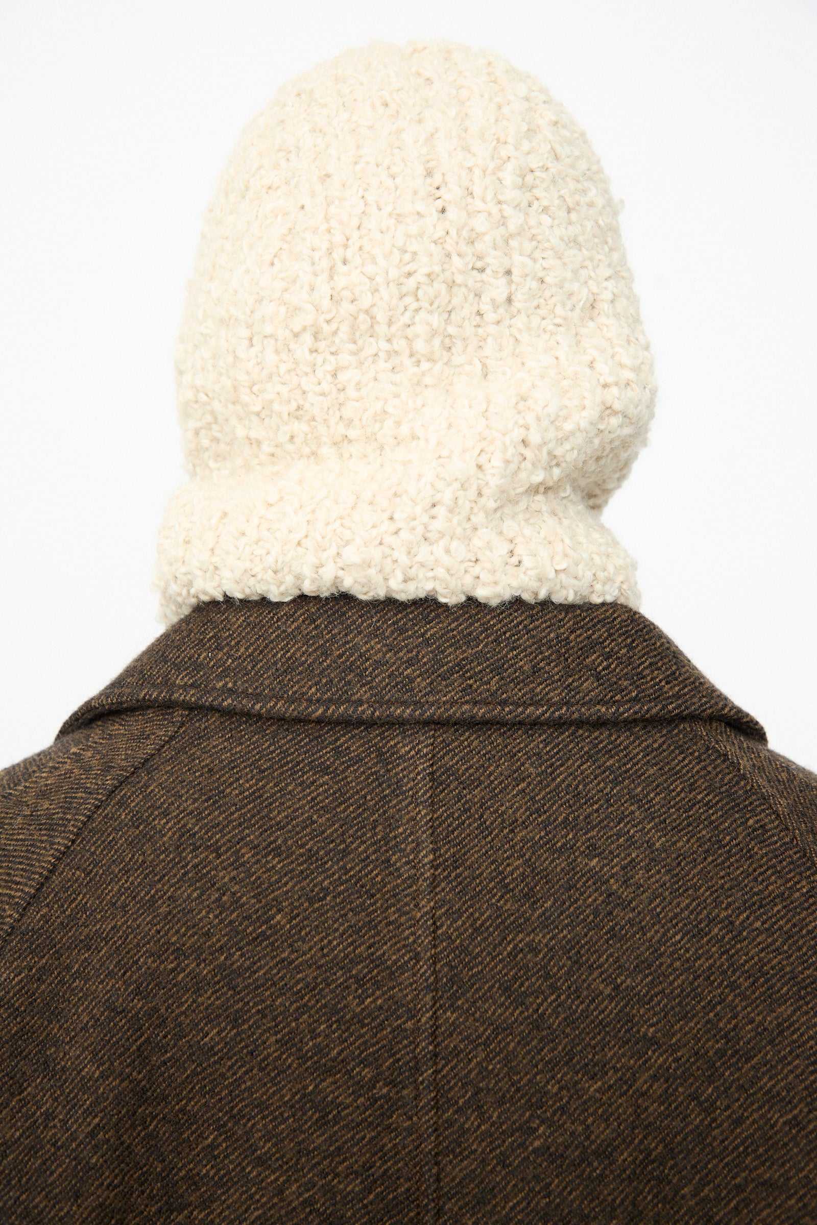 A person wearing a brown wool alpaca blend coat and the SONO Bruna Balaclava in Natural, featuring a thick, cream-colored knit with a subtle ribbed texture, photographed from behind against a plain light background.