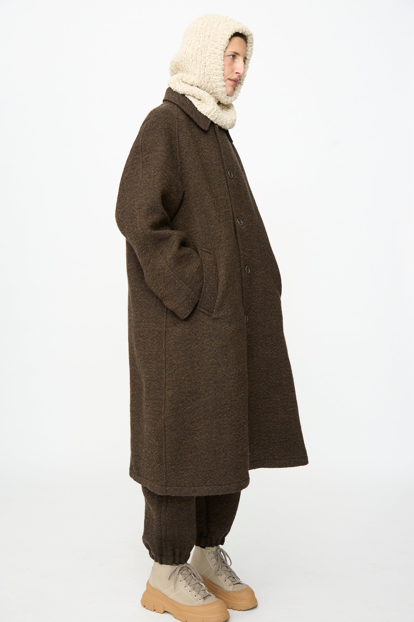 A person stands in profile wearing a long brown coat and matching pants, complemented by the SONO Bruna Balaclava in Natural—a handknit beige wool-alpaca blend with a subtle ribbed texture—and paired with beige boots, set against a plain white background.