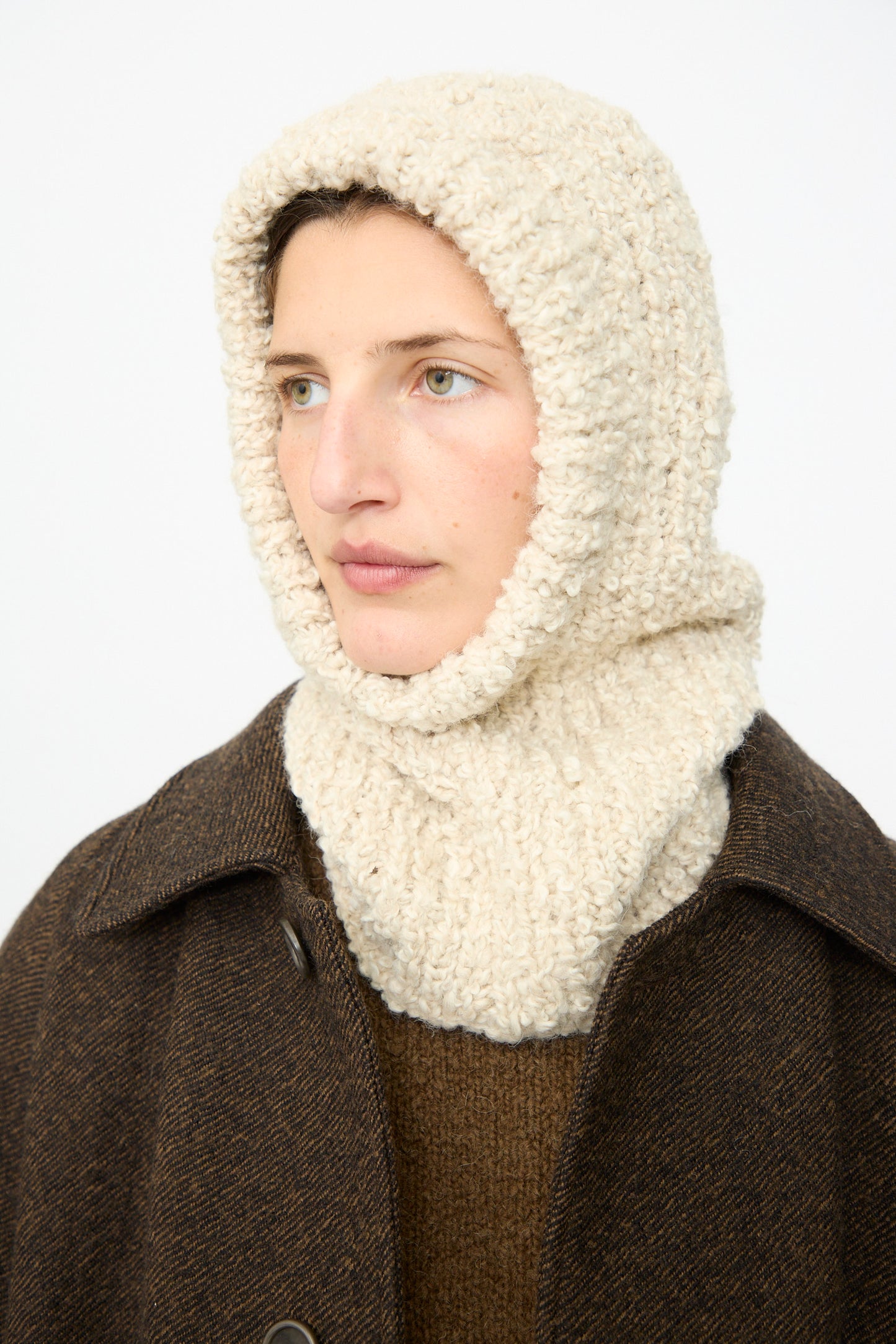 Wearing the SONO Bruna Balaclava in Natural—a handknit, textured cream knit hood and scarf—the person pairs it with a brown coat and a ribbed brown sweater, gazing slightly to the side against a plain white background.