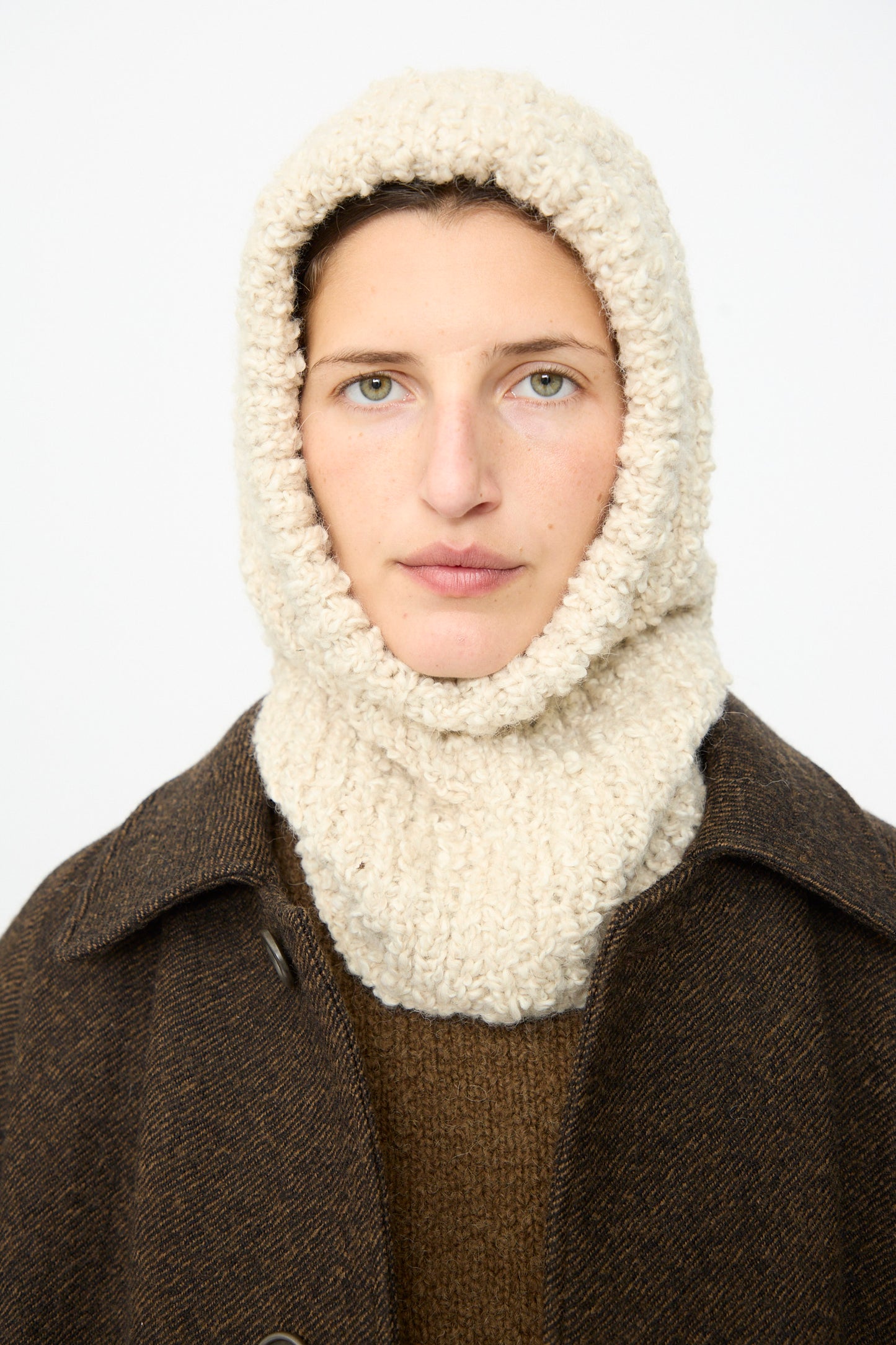 A person wearing the Bruna Balaclava in Natural by SONO—a cream-colored, handknit balaclava featuring a ribbed texture—paired with a brown coat and looking directly at the camera against a plain white background.
