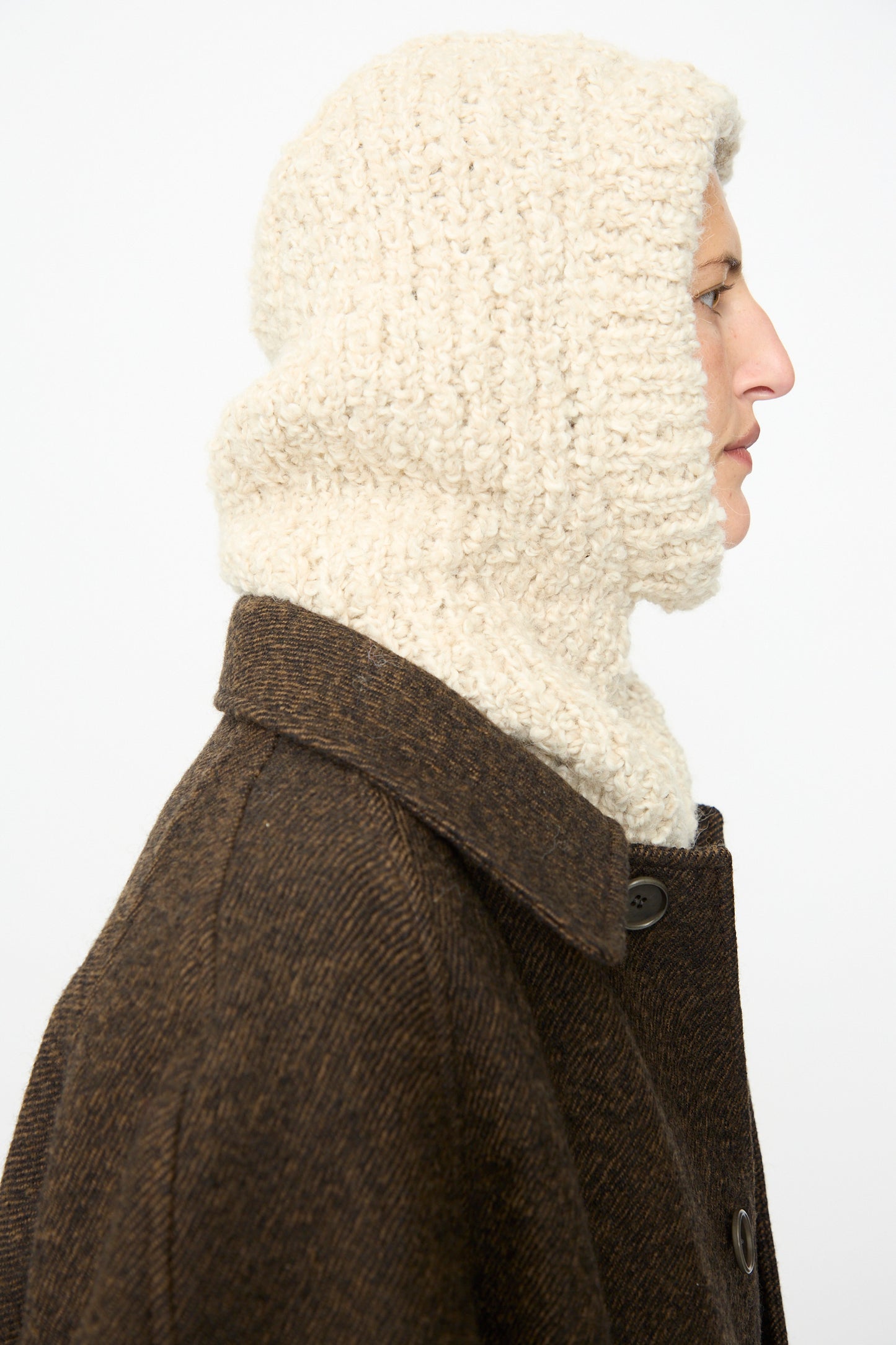 A person in profile wears the Bruna Balaclava in Natural by SONO, featuring a handknit ribbed texture in cream, along with a dark brown buttoned coat against a plain light background.