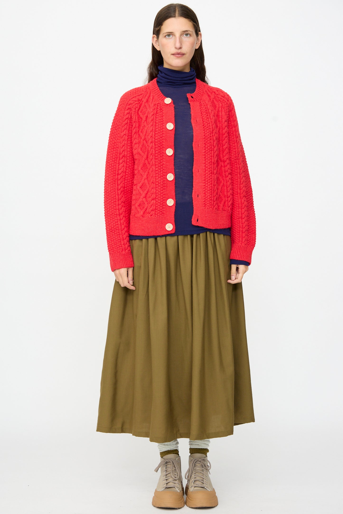 A woman stands against a white background wearing the SONO Candy Cardigan in Red, paired with a navy turtleneck, olive green skirt, olive leggings, and tan lace-up shoes.