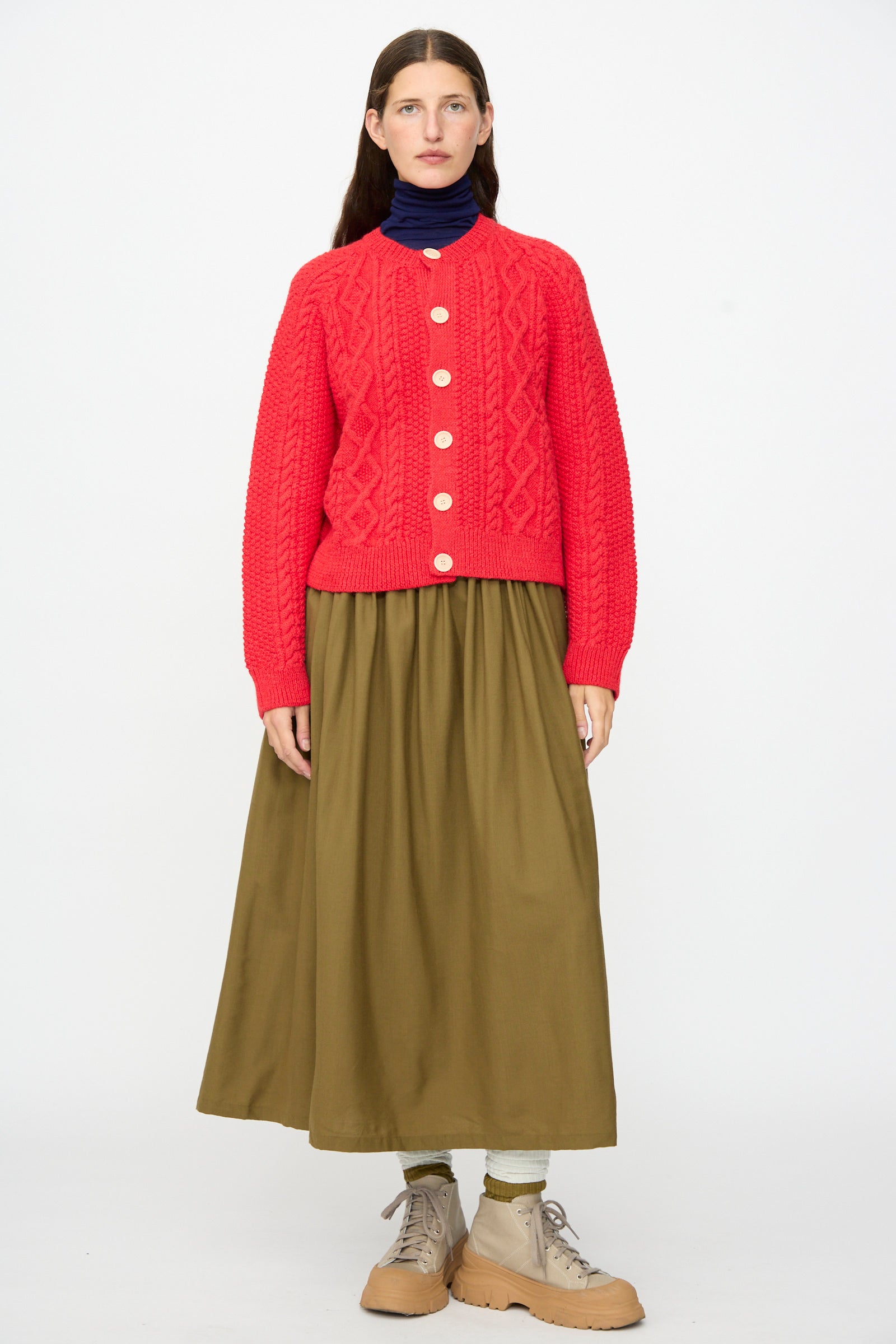 A person stands against a white background wearing the SONO Candy Cardigan in Red, paired with an olive green long skirt, beige boots, and a navy turtleneck.