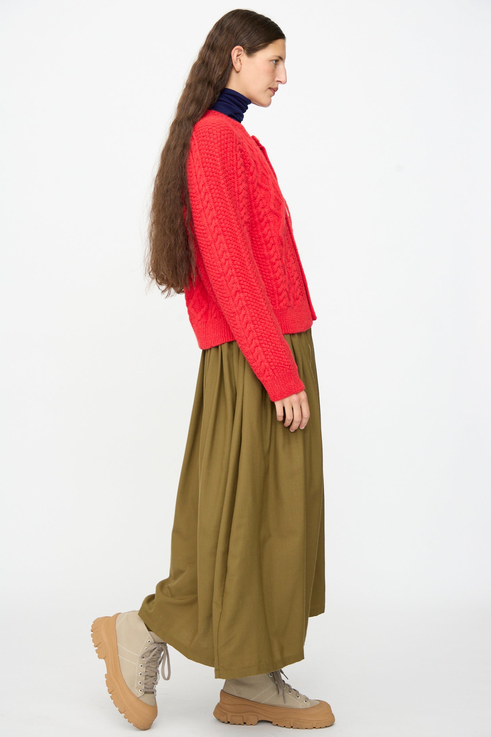 Wearing the SONO Candy Cardigan in Red, a UK-made wool alpaca piece, this person is shown in profile paired with an olive green long skirt, beige lace-up boots, and a dark turtleneck, standing against a plain white background.