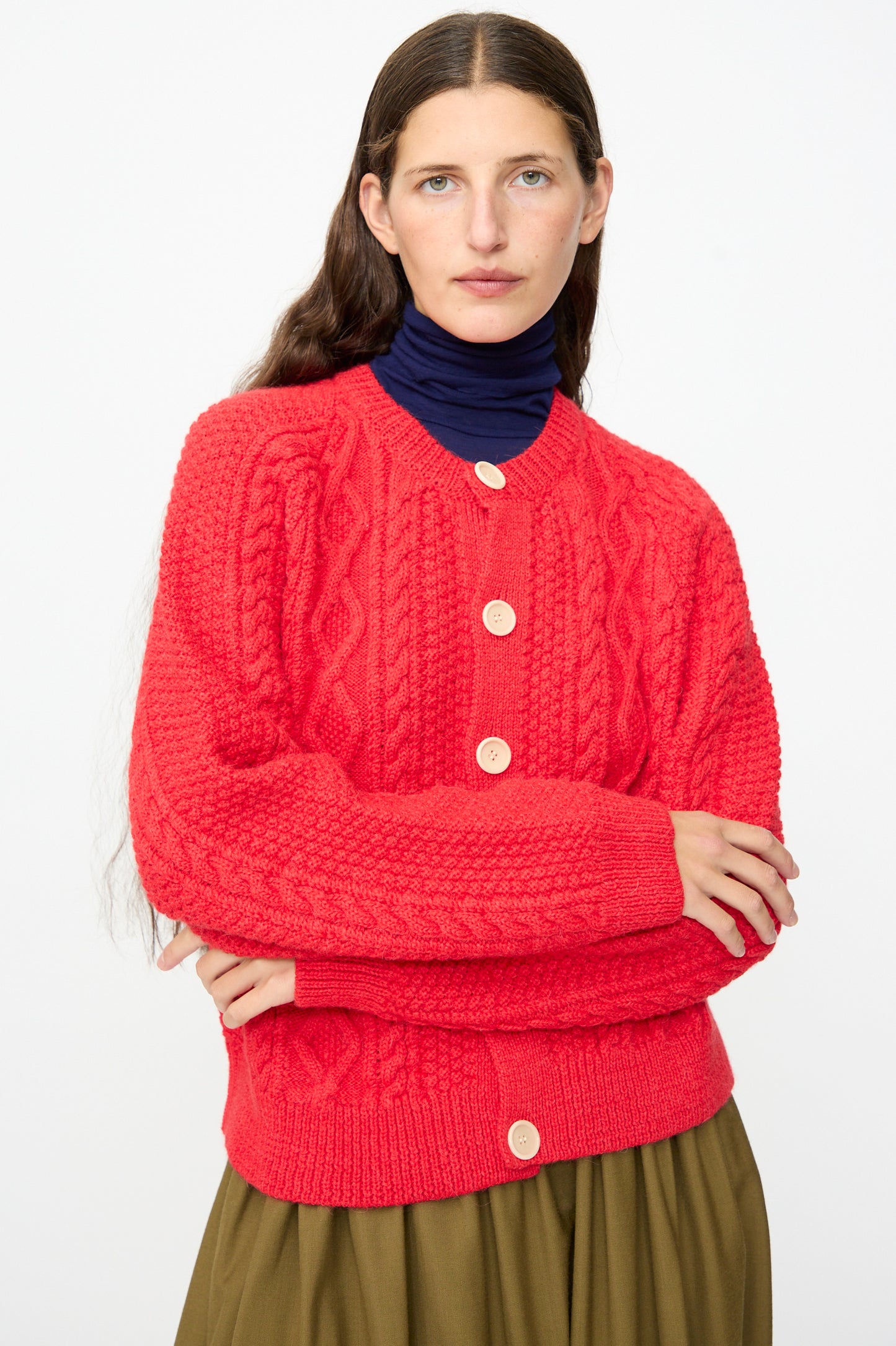 Wearing SONO’s Candy Cardigan in Red—a bright wool alpaca crewneck with large buttons, made in the UK—a person with long brown hair pairs it with a navy turtleneck and an olive green skirt while standing against a plain white background.