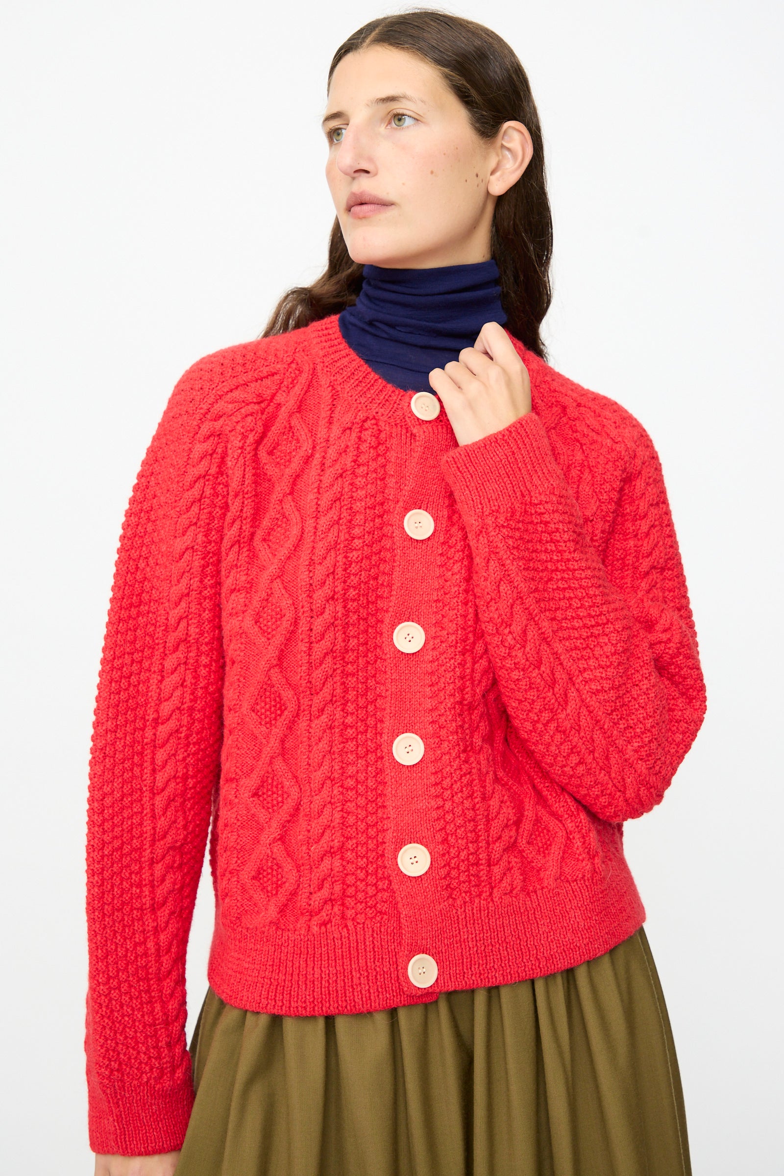 Wearing the SONO Candy Cardigan in Red—a vibrant wool-alpaca crewneck cardigan made in the UK, featuring large white buttons—layered over a navy turtleneck and paired with an olive green skirt, this person stands against a plain light background.