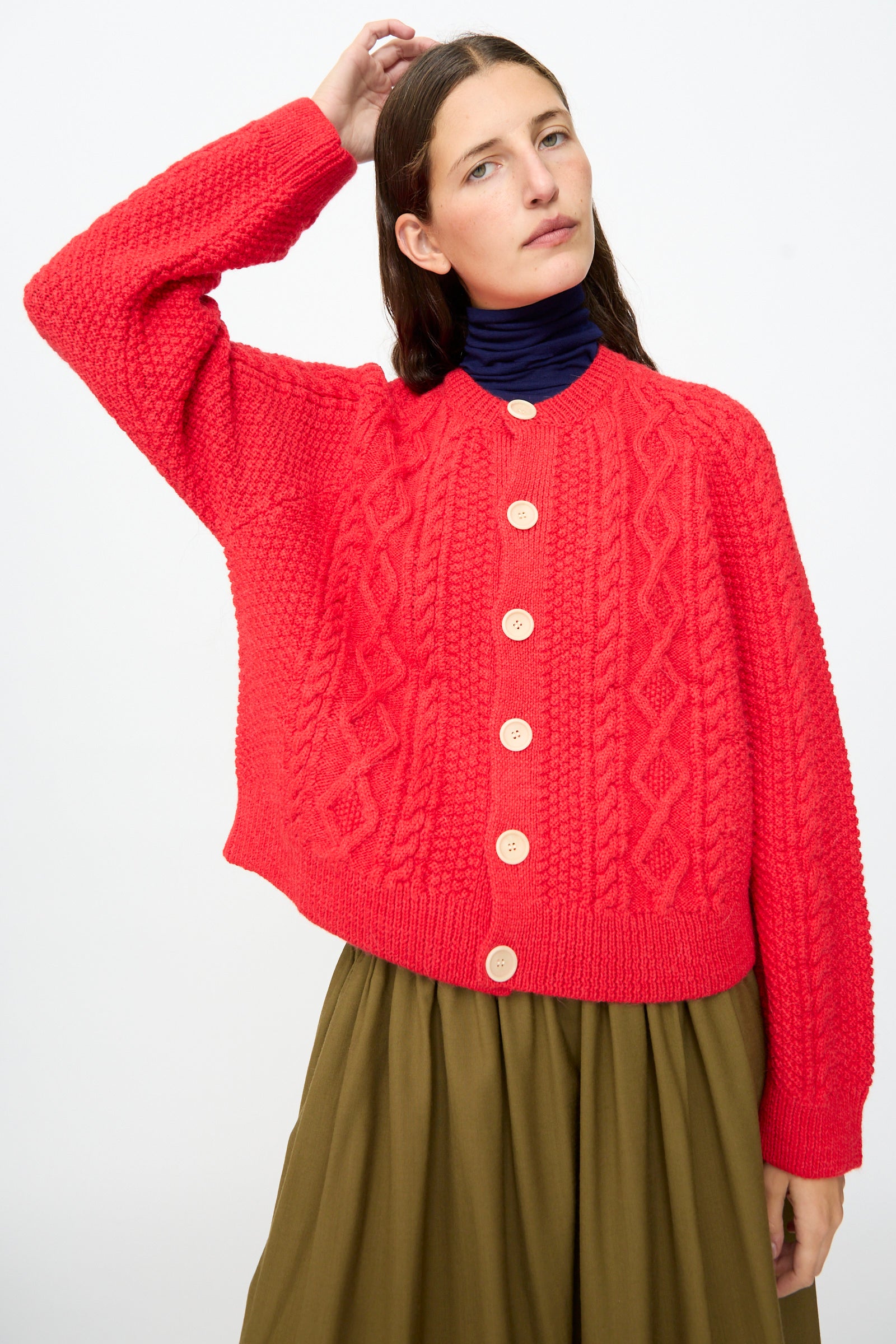 Wearing the SONO Candy Cardigan in Red, styled as a button-up over a dark turtleneck and olive green skirt, a person poses with one hand raised behind their head against a plain white background.