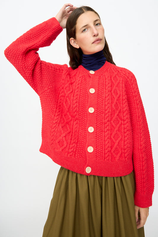 Wearing the SONO Candy Cardigan in Red, styled as a button-up over a dark turtleneck and olive green skirt, a person poses with one hand raised behind their head against a plain white background.