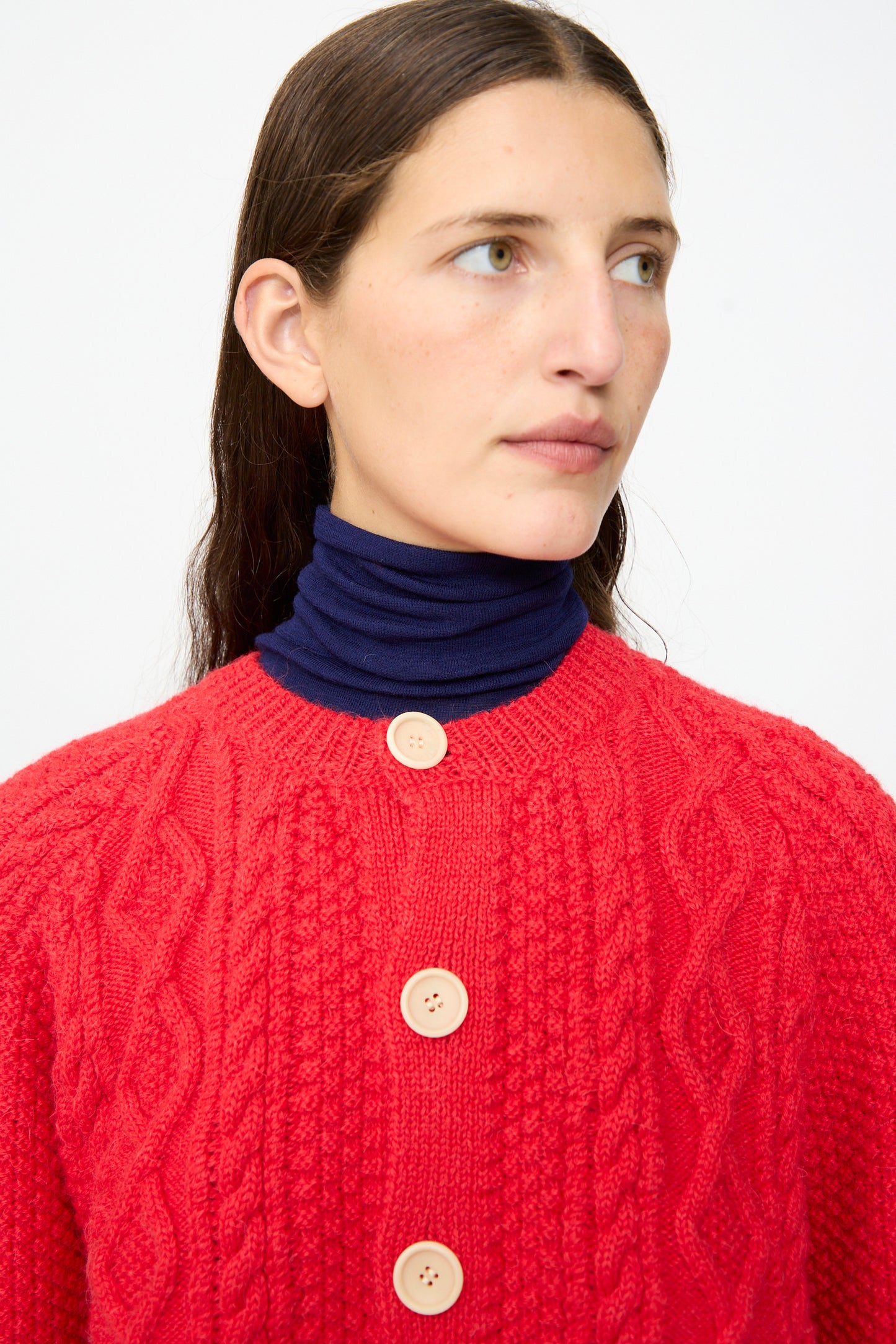 A person wearing the SONO Candy Cardigan in Red, a cable-knit wool alpaca sweater with large buttons, layered over a navy blue turtleneck and gazing slightly to the side against a plain background.