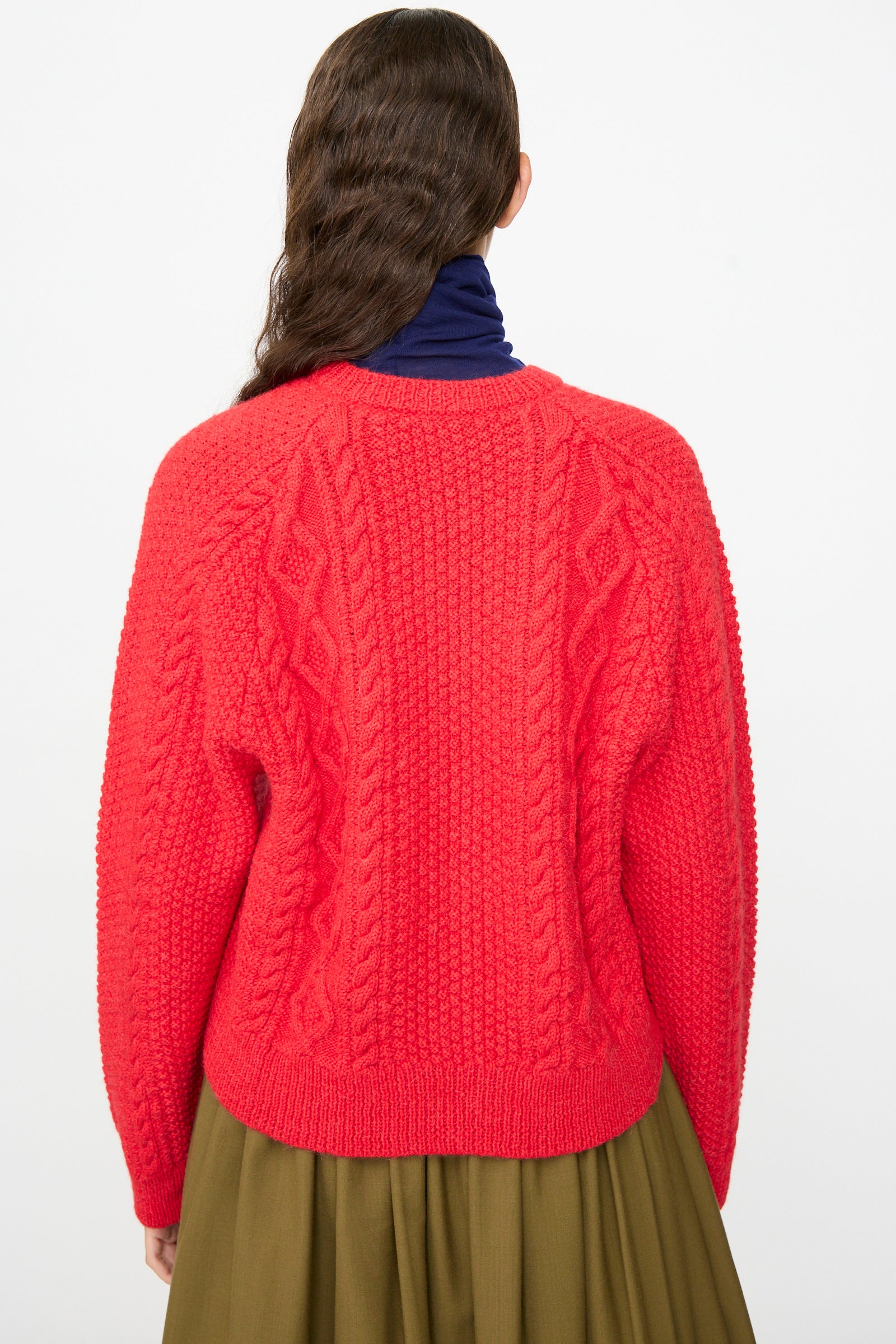 Person with long wavy brown hair, shown from the back against a plain background, wearing the SONO Candy Cardigan in Red—a textured wool alpaca cardigan made in the UK—layered over a dark blue turtleneck and an olive green skirt.