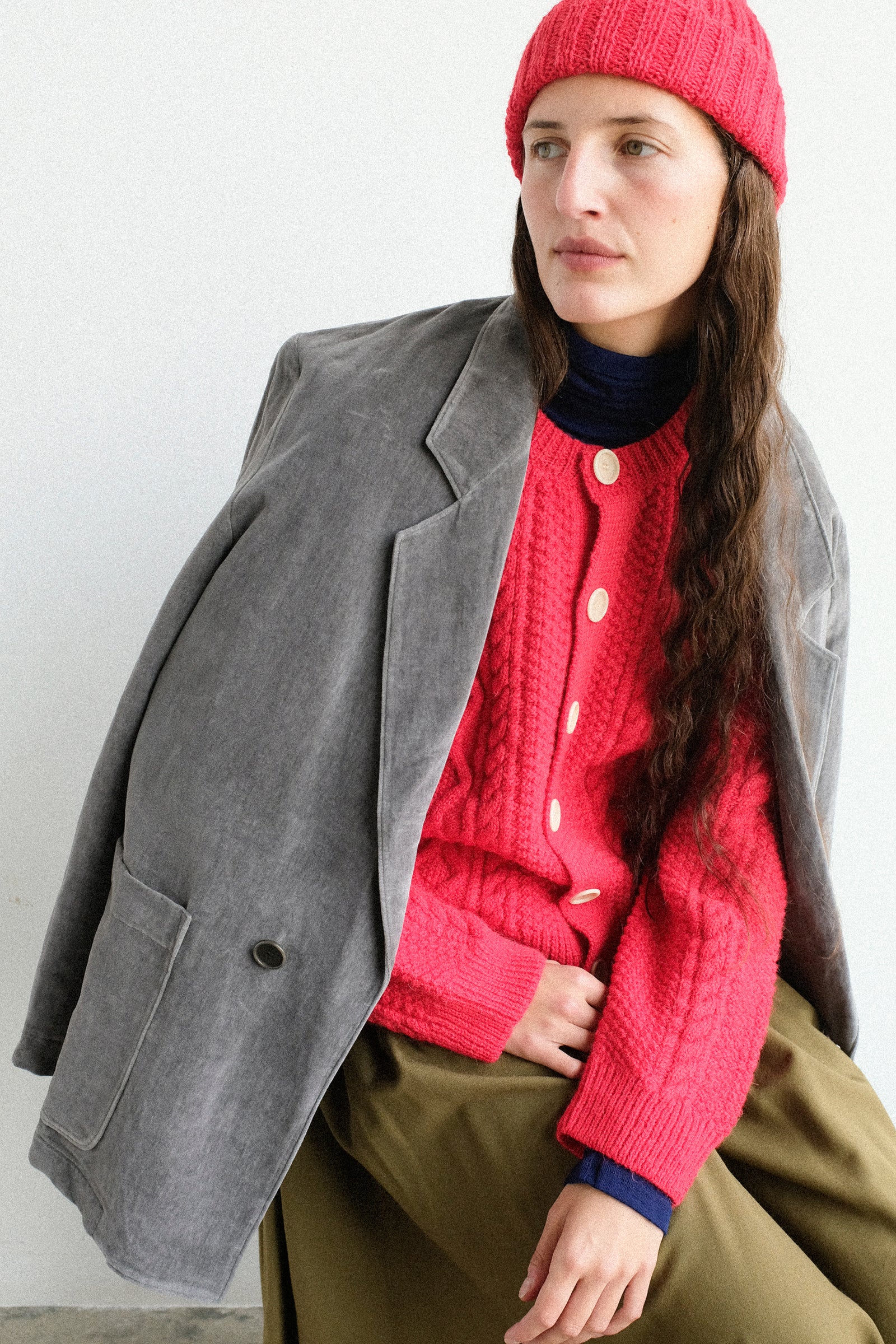 A woman wearing a SONO's Gyu Beanie, Candy Cardigan, Juri Jacket, and Skye Skirt, posed against a plain light gray wall.