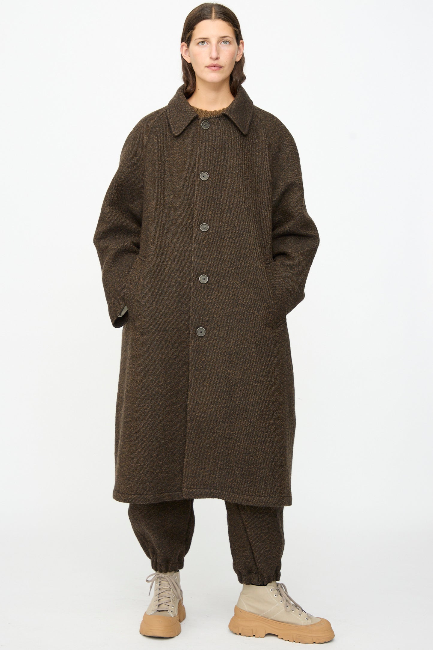 A person wearing the SONO Carl Coat in Blackmud—an oversized long wool coat made in London—over matching brown pants and tan lace-up shoes, standing against a plain white background.