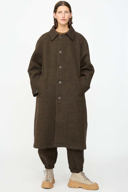 A person wearing the SONO Carl Coat in Blackmud—an oversized long wool coat made in London—over matching brown pants and tan lace-up shoes, standing against a plain white background.
