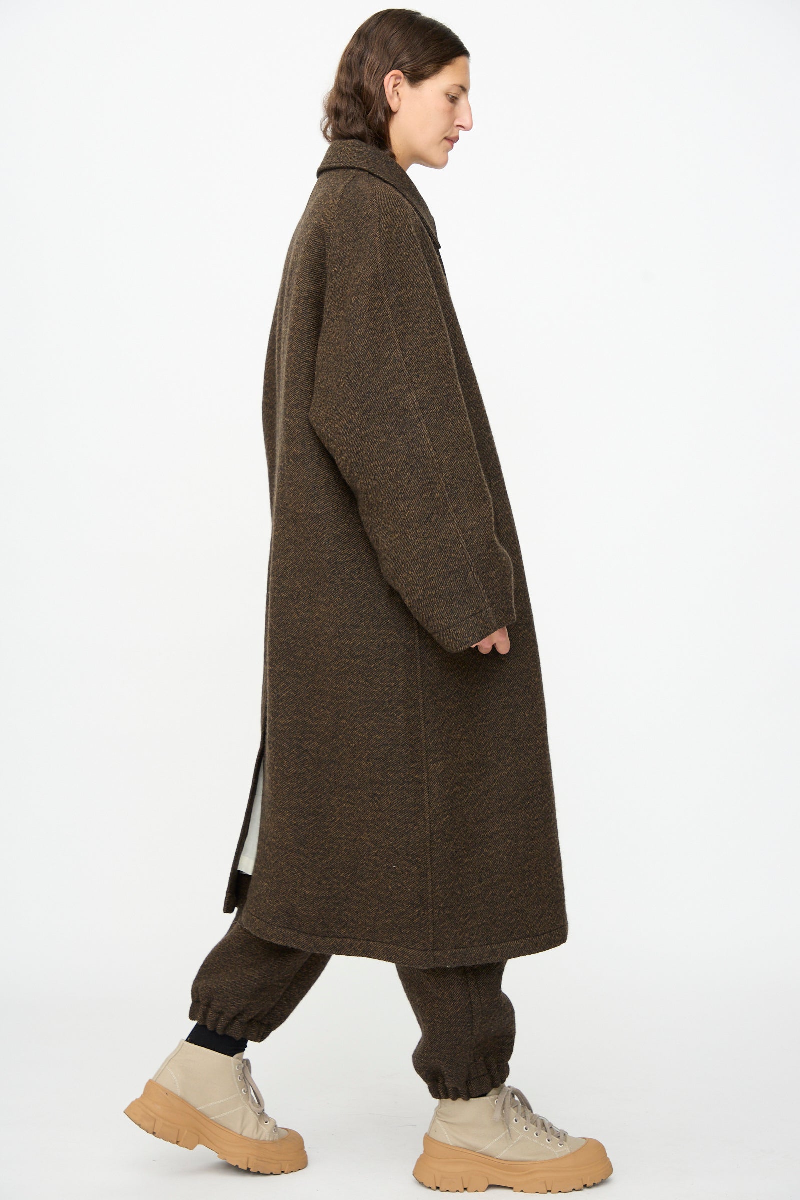 A person stands in profile against a plain white background, wearing the SONO Carl Coat in Blackmud with matching pants and light beige boots.