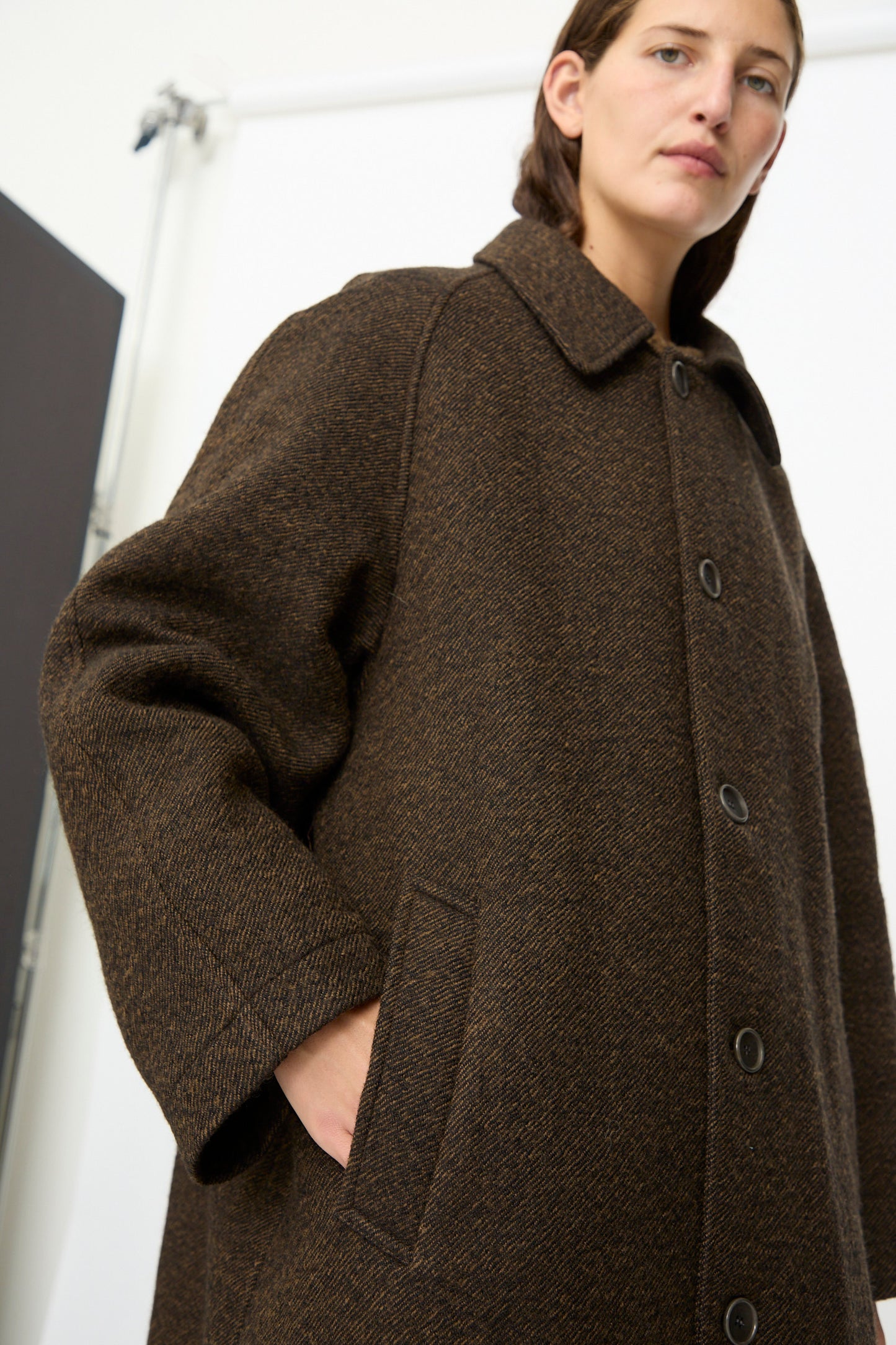 A person wearing the SONO Carl Coat in Blackmud—an oversized, long, brown textured wool coat with buttons and pockets made in London—stands indoors with one hand in the pocket, looking slightly down at the camera.