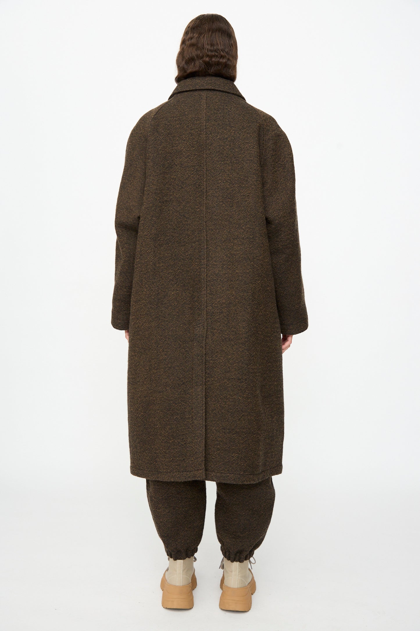 A person stands with their back to the camera, wearing the oversized SONO Carl Coat in Blackmud, paired with brown pants and beige shoes against a plain white background.