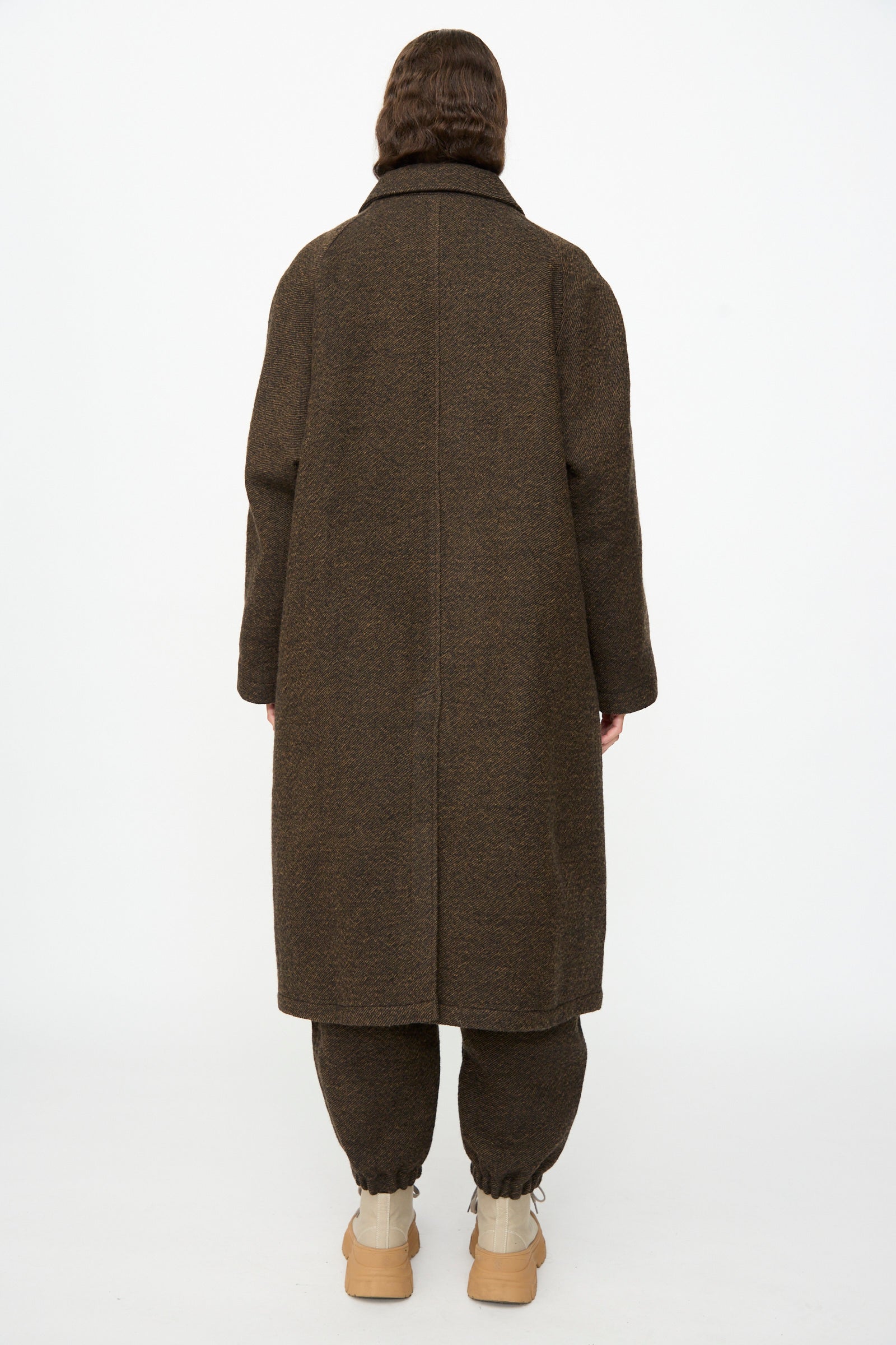 A person stands with their back to the camera, wearing the oversized SONO Carl Coat in Blackmud, paired with brown pants and beige shoes against a plain white background.