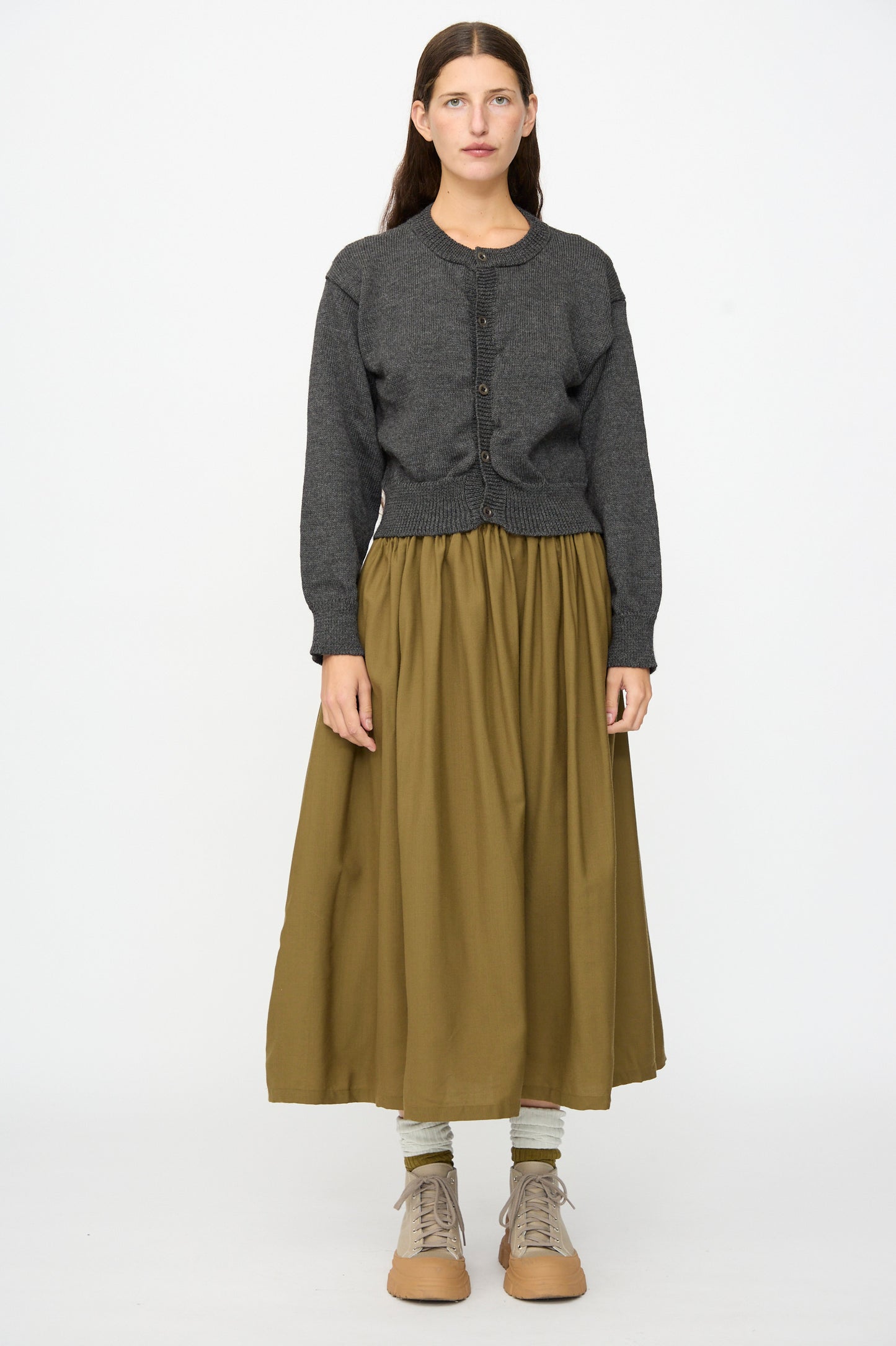 A woman stands against a plain white background, wearing the Caspa Cardigan in Charcoal by SONO—a dark gray wool alpaca crewneck cardigan made in the UK—paired with an olive green ankle-length skirt, light socks, and tan lace-up shoes.