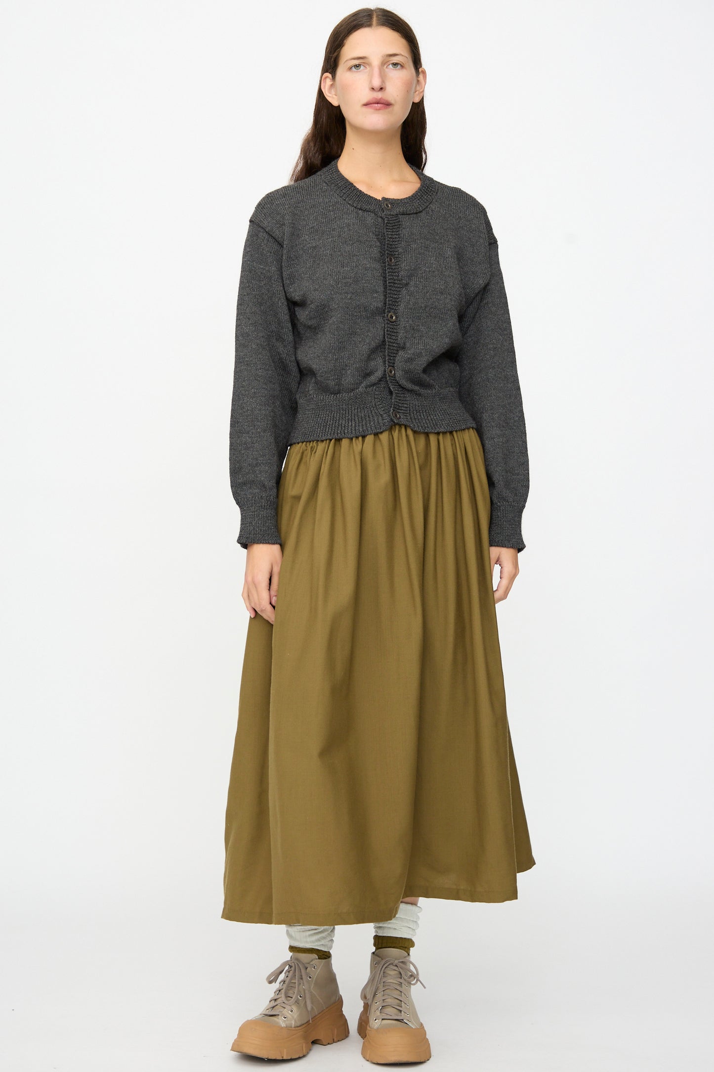 A person stands facing forward against a plain white background, wearing the SONO Caspa Cardigan in Charcoal—made in the UK—a cozy olive green long skirt, light leggings, and tan lace-up boots.