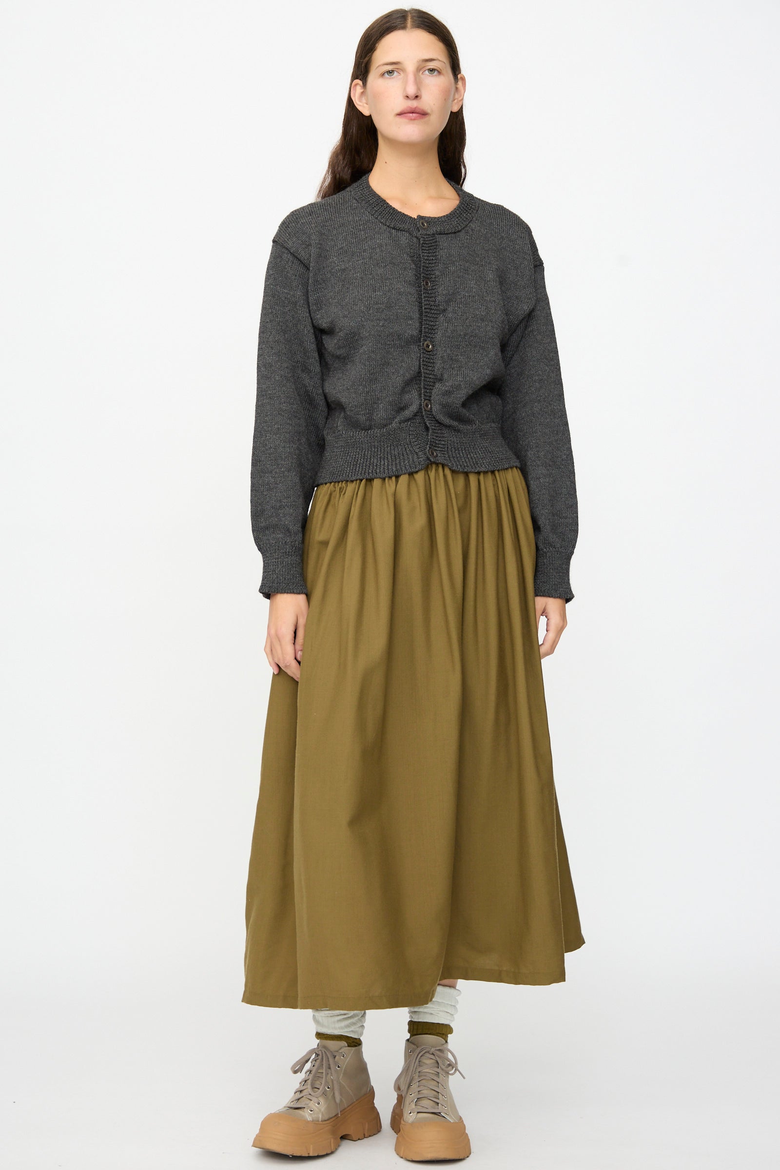 A person stands facing forward against a plain white background, wearing the SONO Caspa Cardigan in Charcoal—made in the UK—a cozy olive green long skirt, light leggings, and tan lace-up boots.