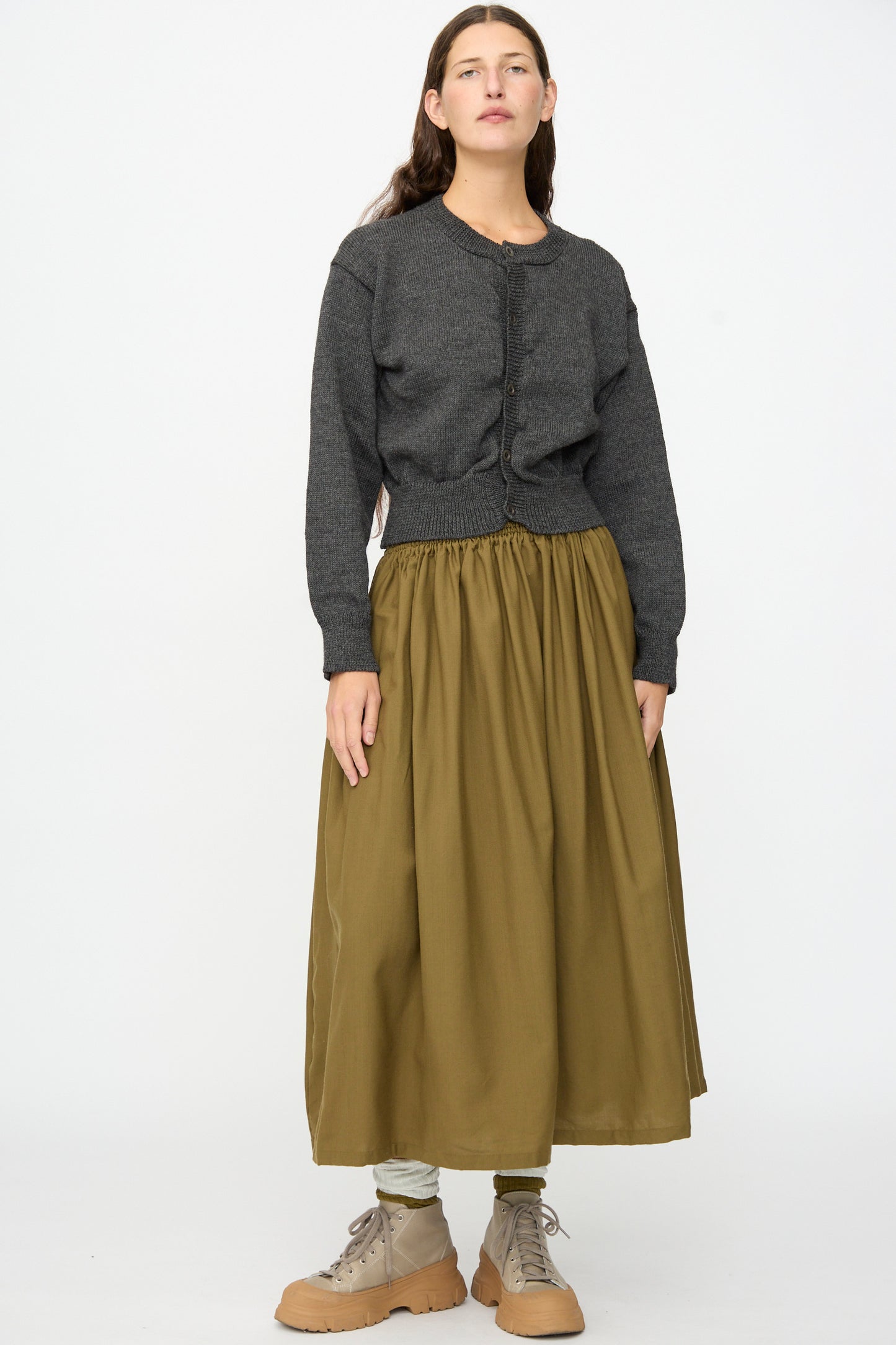 A person stands facing forward against a plain white background, wearing the SONO Caspa Cardigan in Charcoal, an olive green ankle-length skirt, beige lace-up boots, and light-colored leggings.
