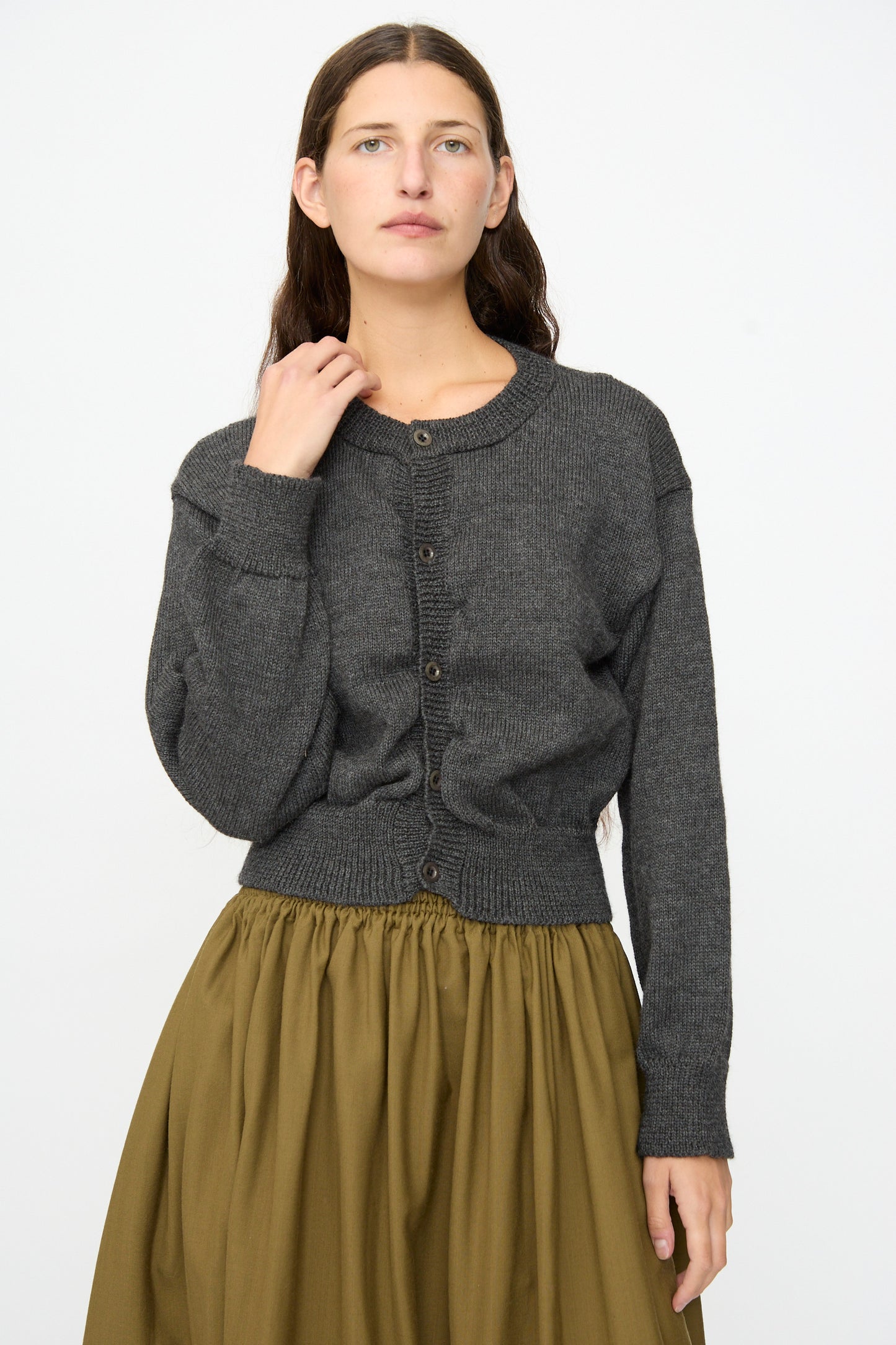 A woman with long brown hair wears the Caspa Cardigan in Charcoal by SONO, made from wool alpaca in the UK, paired with an olive green skirt and standing against a plain white background.
